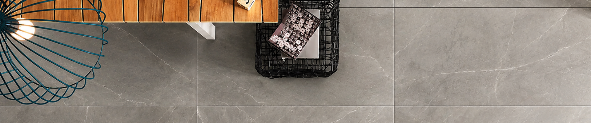 Top-down view of a modern interior space focusing on a gray tiled floor, part of a wooden table, and a black wire basket with magazines inside.