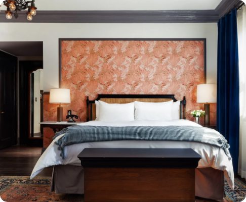 Hotel bedroom with a bed, white pillows, gray blanket, blue drapes, two lamps, and a floral wall behind the bed.