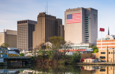 City skyline with several tall buildings, one featuring an American flag on its facade, and a body of water reflecting the cityscape in the foreground.
