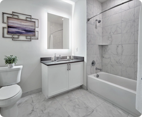 A modern bathroom featuring a bathtub in a tiled shower area, a white vanity with a dark countertop, a large mirror, a toilet, and minimalist wall decor with a purple landscape art piece.