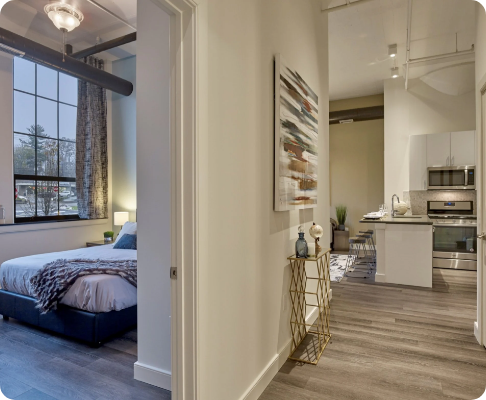 Interior view of a modern apartment with a bedroom on the left, featuring a bed with a gray blanket, window with curtains, and a side table with a lamp, and a kitchen on the right with white cabinets, stainless steel appliances, and a wooden floor.