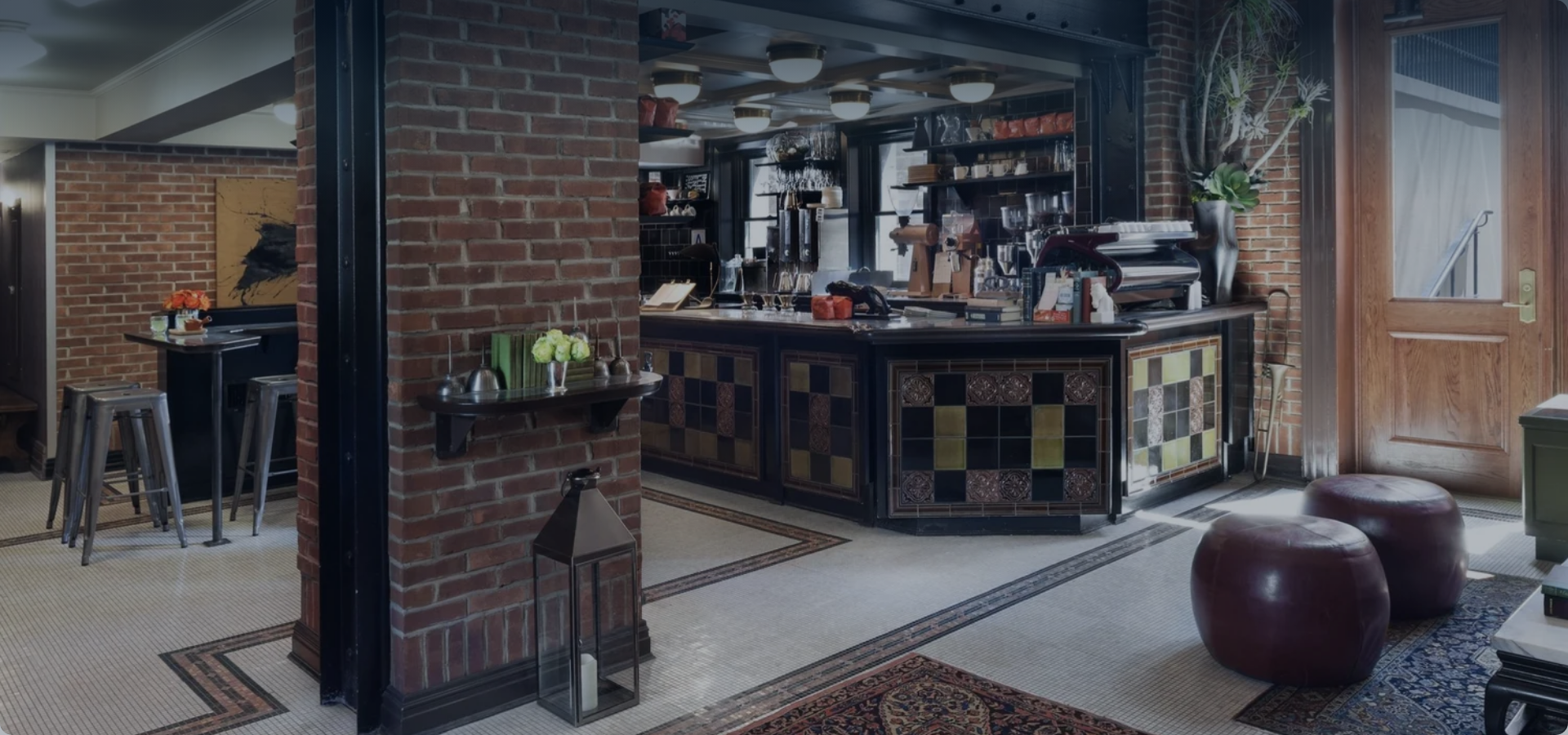 Interior view of a cozy restaurant or cafe featuring a brick wall, a bar counter with colorful tiles, and a table with chairs. Decor includes flowers, artwork, and natural lighting from the door window.