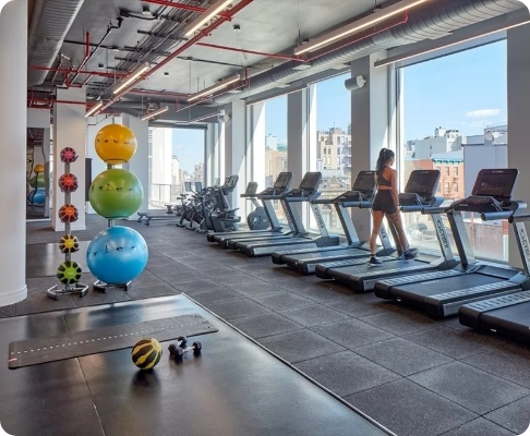 Modern gym with treadmills by large windows, stacked colorful exercise balls, and workout equipment, with a woman using a treadmill.