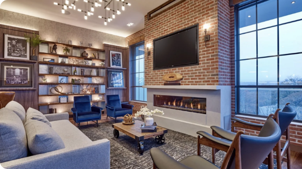 Living room with brick walls, a large flat-screen TV above a modern fireplace, a gray sofa, blue armchairs, wooden shelves with decorative items, a large window, and contemporary lighting.