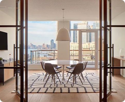 Modern dining room with a round table, four chairs, and a large pendant light, featuring floor-to-ceiling windows with city skyline view.