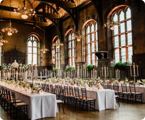 Elegant banquet hall with long tables decorated with white tablecloths, candlesticks, and floral centerpieces, featuring large arched windows and chandeliers.