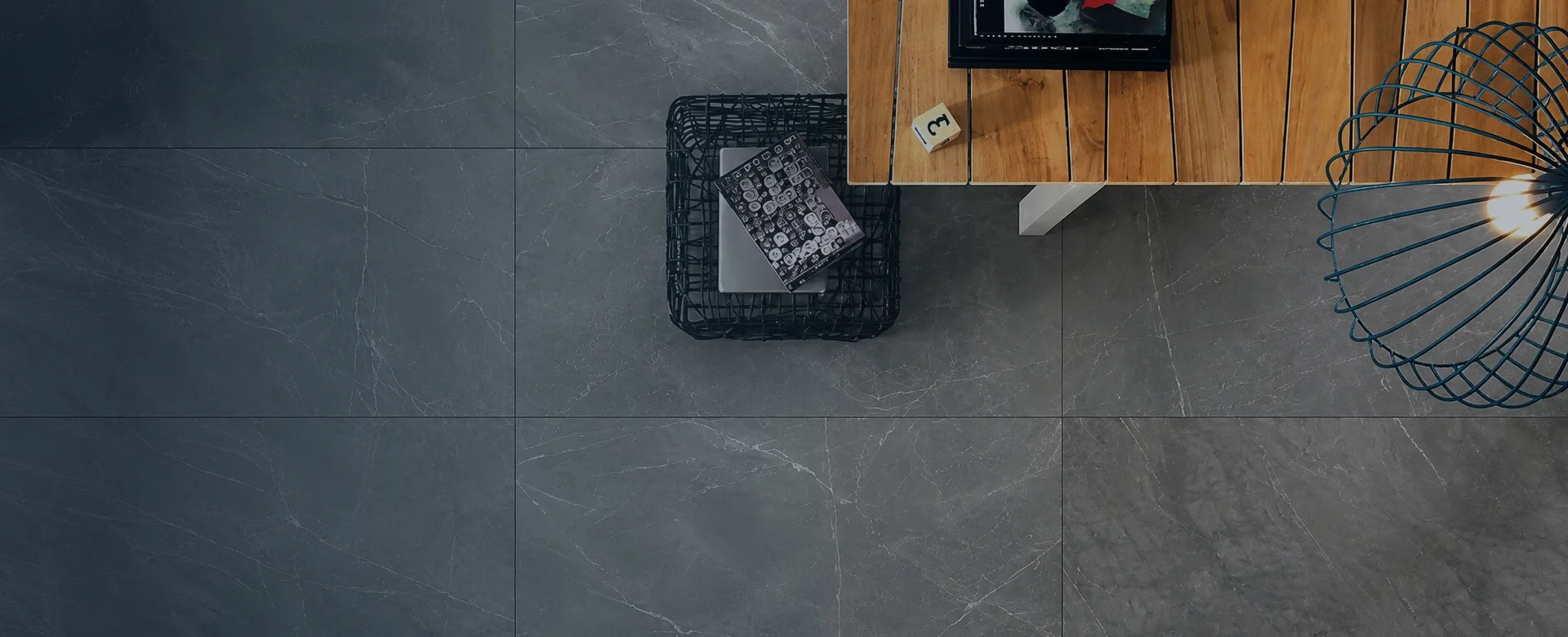 Top-down view of a modern interior with gray tiled flooring, a black wire basket containing a magazine and laptop, a wooden table with a frame and a cube block, and a black wire pendant light.