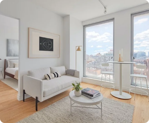 Modern living room with white sofa, coffee table, large window overlooking city buildings, and small dining area with two chairs and a table.