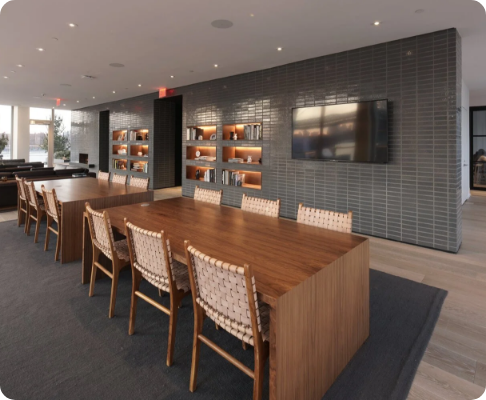 Modern conference room with a large wooden table, woven chairs, a wall-mounted television, built-in shelves with decorative items, and large windows allowing natural light.