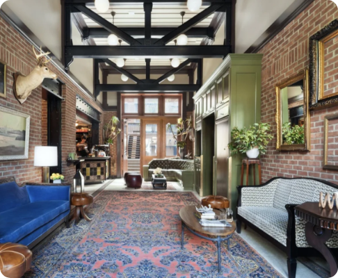 Eclectic living room with brick walls, vintage furniture, and decorative accents, featuring a high ceiling with black structural beams and large windows.