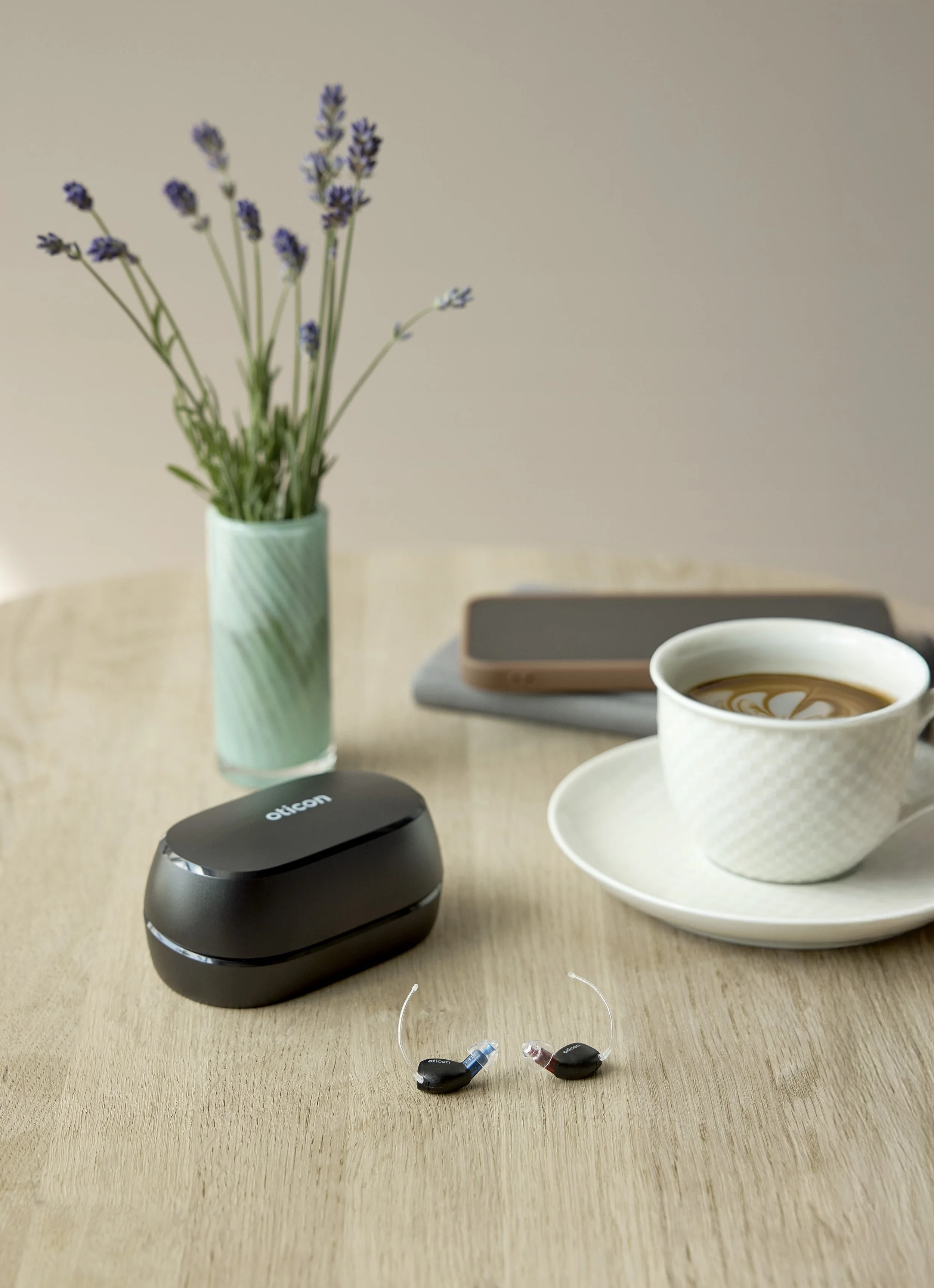 A wooden table with a pair of black Oticon Zeal hearing aids, a black Oticon case, a small white saucer with a cup of coffee, a white mug with latte art, a closed laptop, a gray cloth, a smartphone, and a vase with lavender flowers.