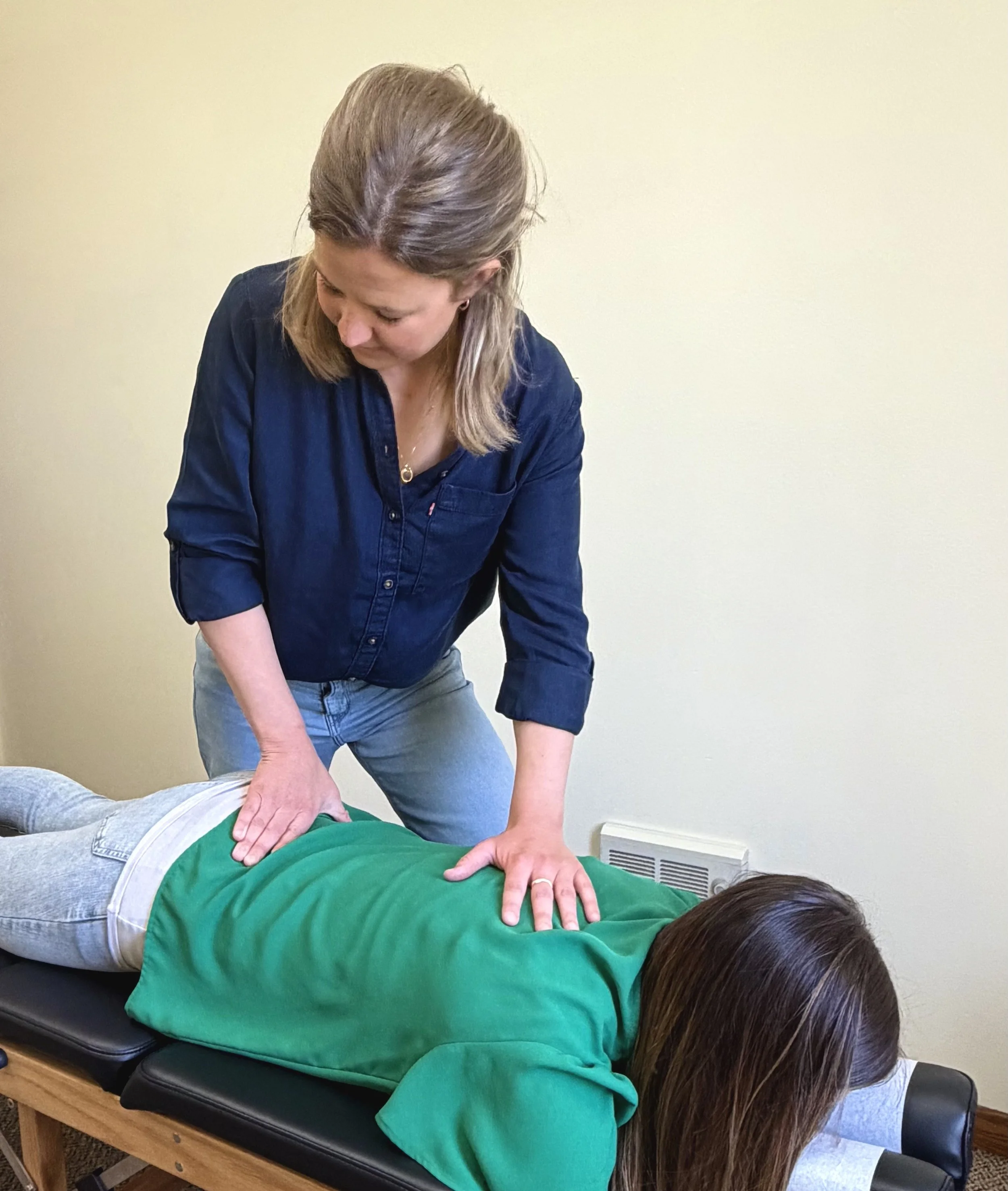 A person in a green sweatshirt lying face down on a treatment table while another person performs a massage or therapy, pressing on their back with both hands.
