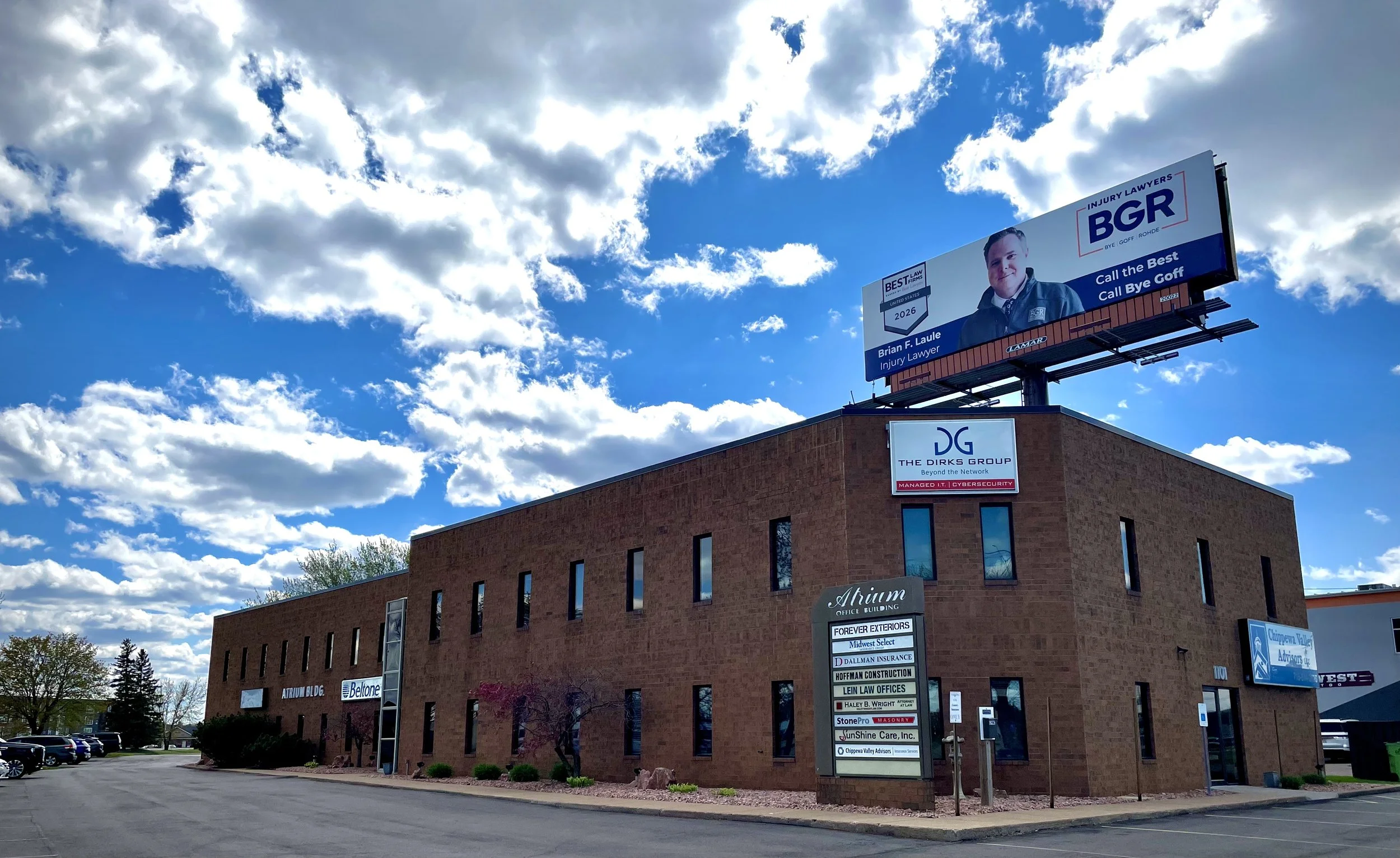 A brick commercial building with multiple signs on it and a parking lot in front, topped with a large billboard promoting injury lawyers with a photo of a man, under a partly cloudy sky.