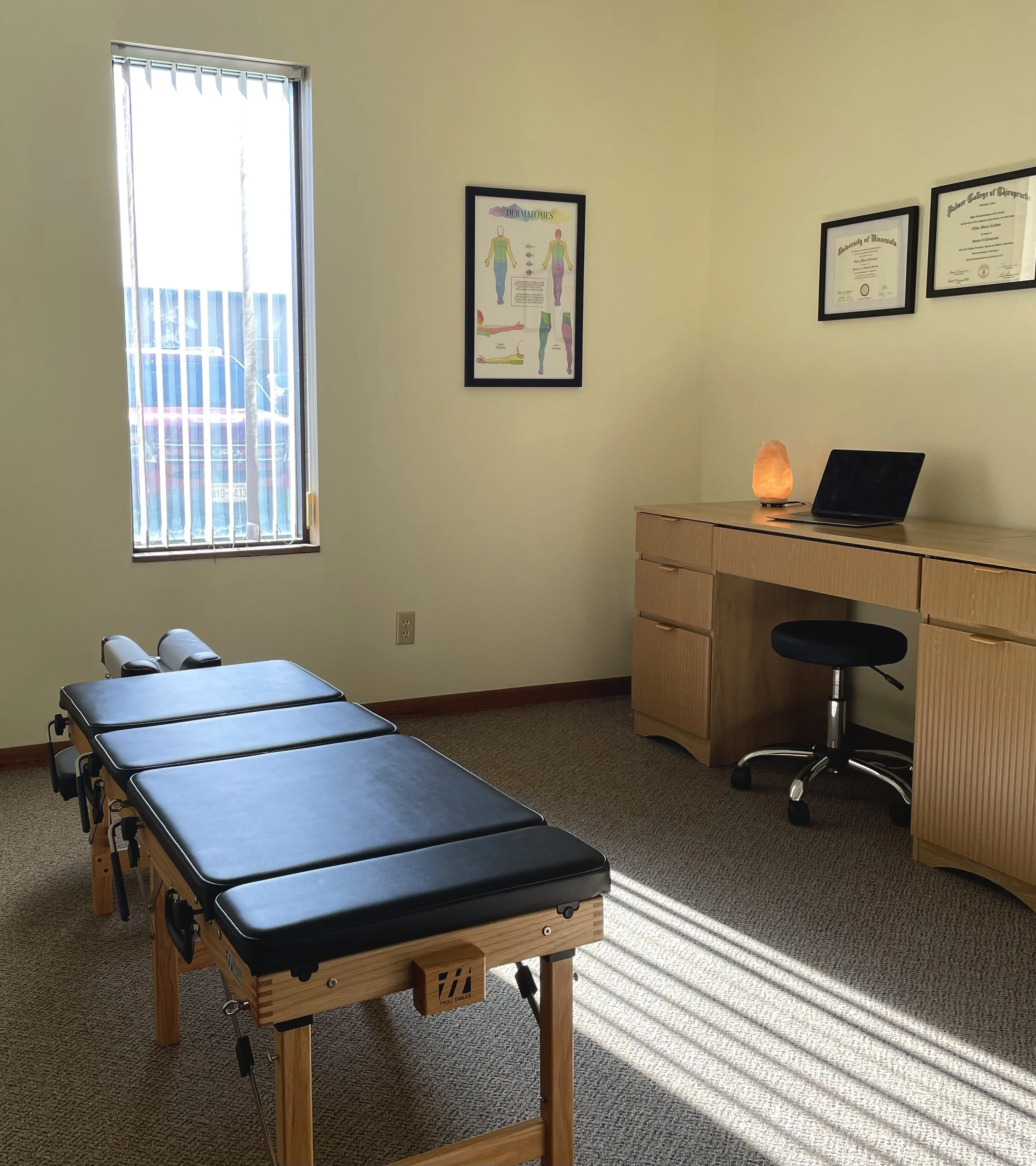 A room with a window with vertical blinds, a massage table, a wooden desk with a lamp, a laptop, and framed certificates and a poster on the wall.