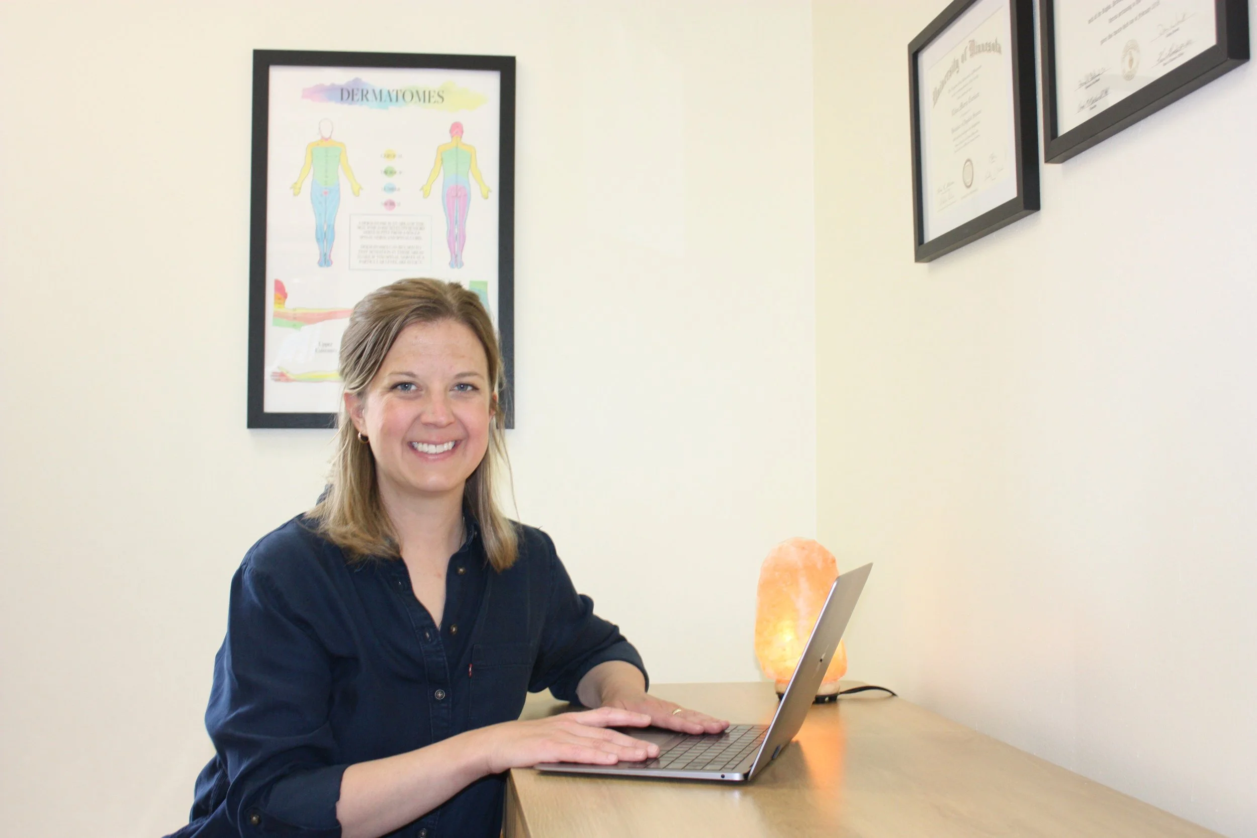 Smiling woman with shoulder-length blonde hair working on a laptop at a wooden desk, with framed diploma and anatomical poster on the wall behind her, and salt lamp on the desk.
