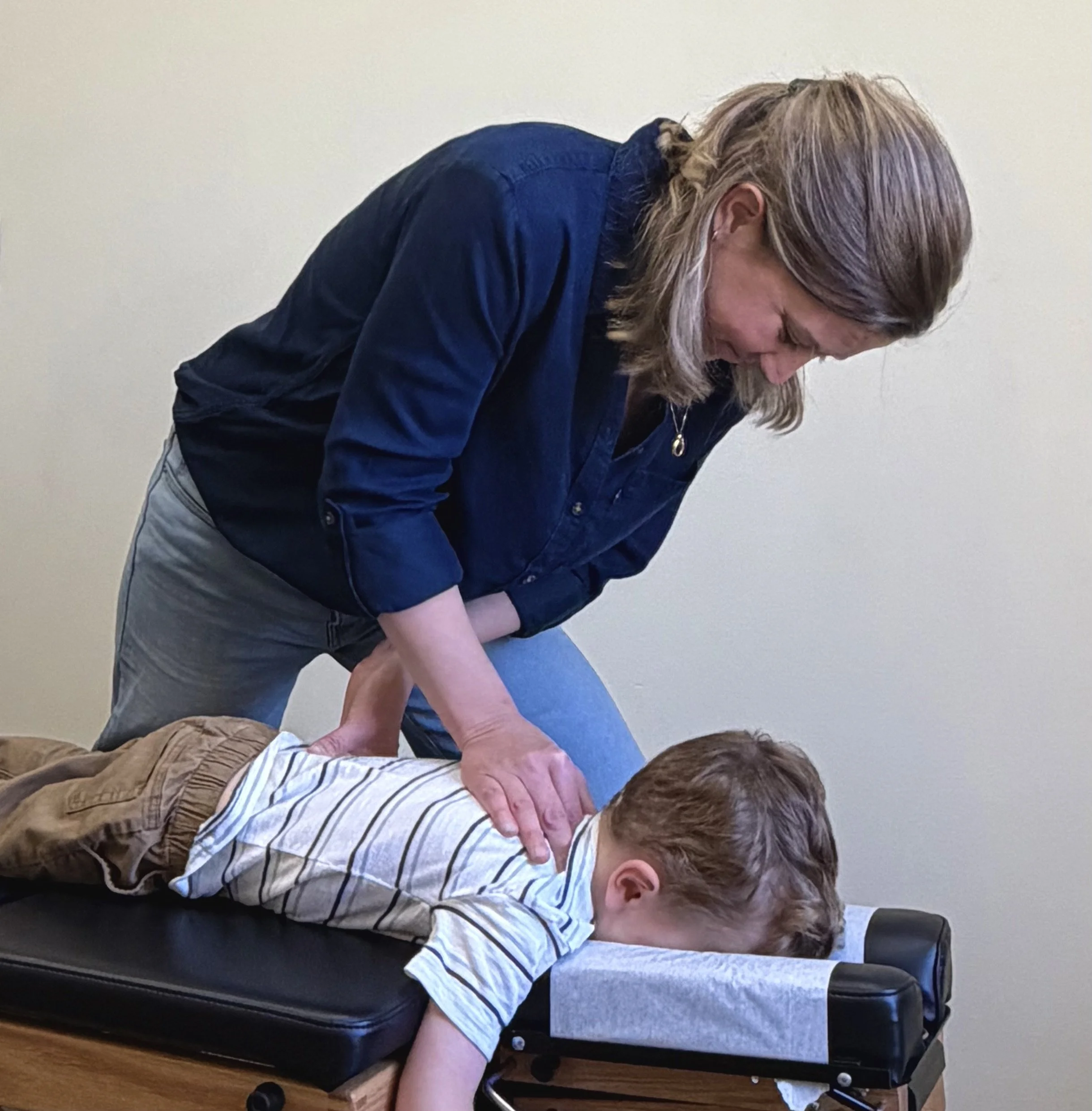 A person receiving CPR training from an instructor in a room with light-colored walls. The person is lying on a training mannequin, and the instructor is practicing chest compressions.
