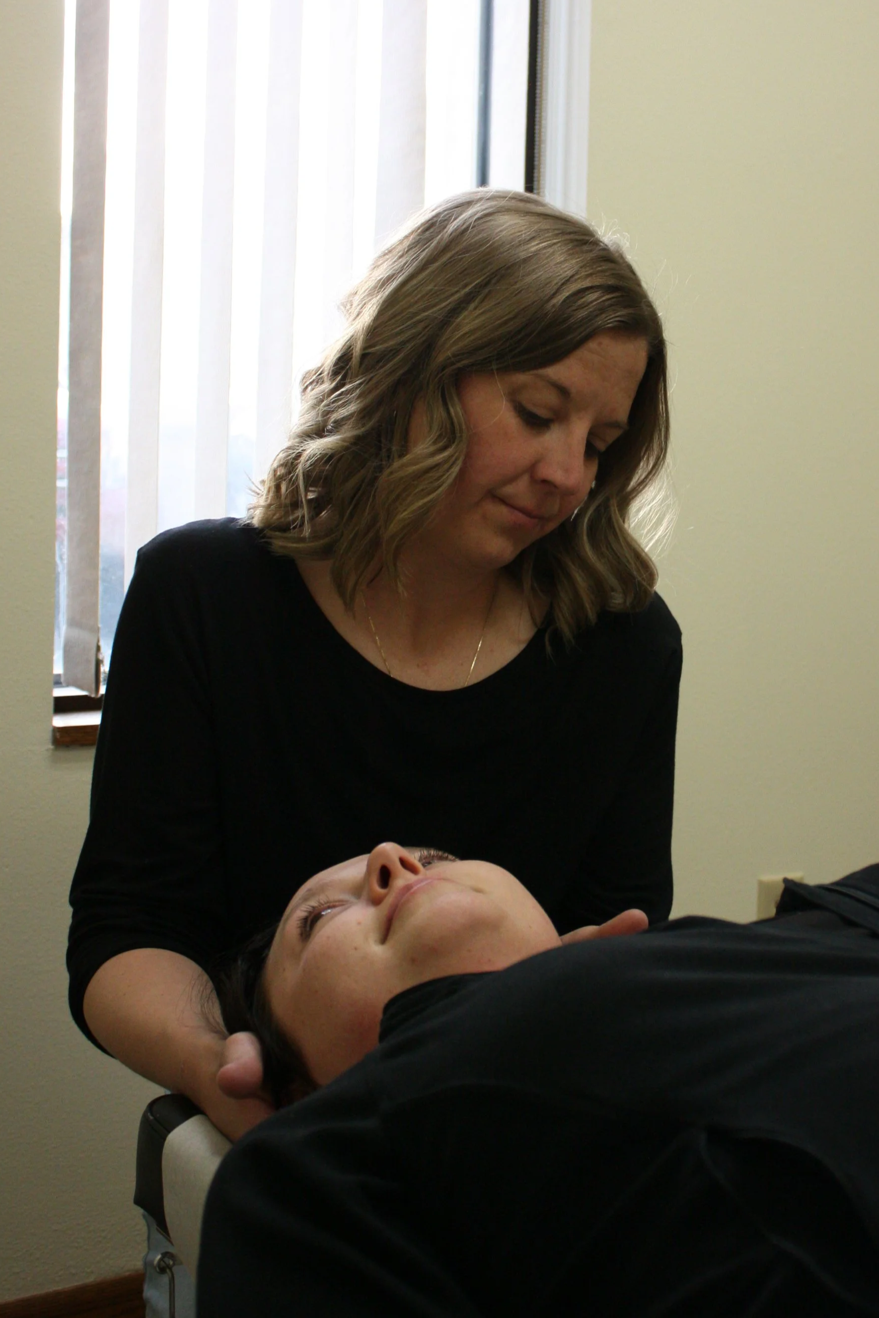 A woman with shoulder-length wavy hair dressed in black is performing acupuncture on a person lying on a treatment table indoors next to a window with vertical blinds.