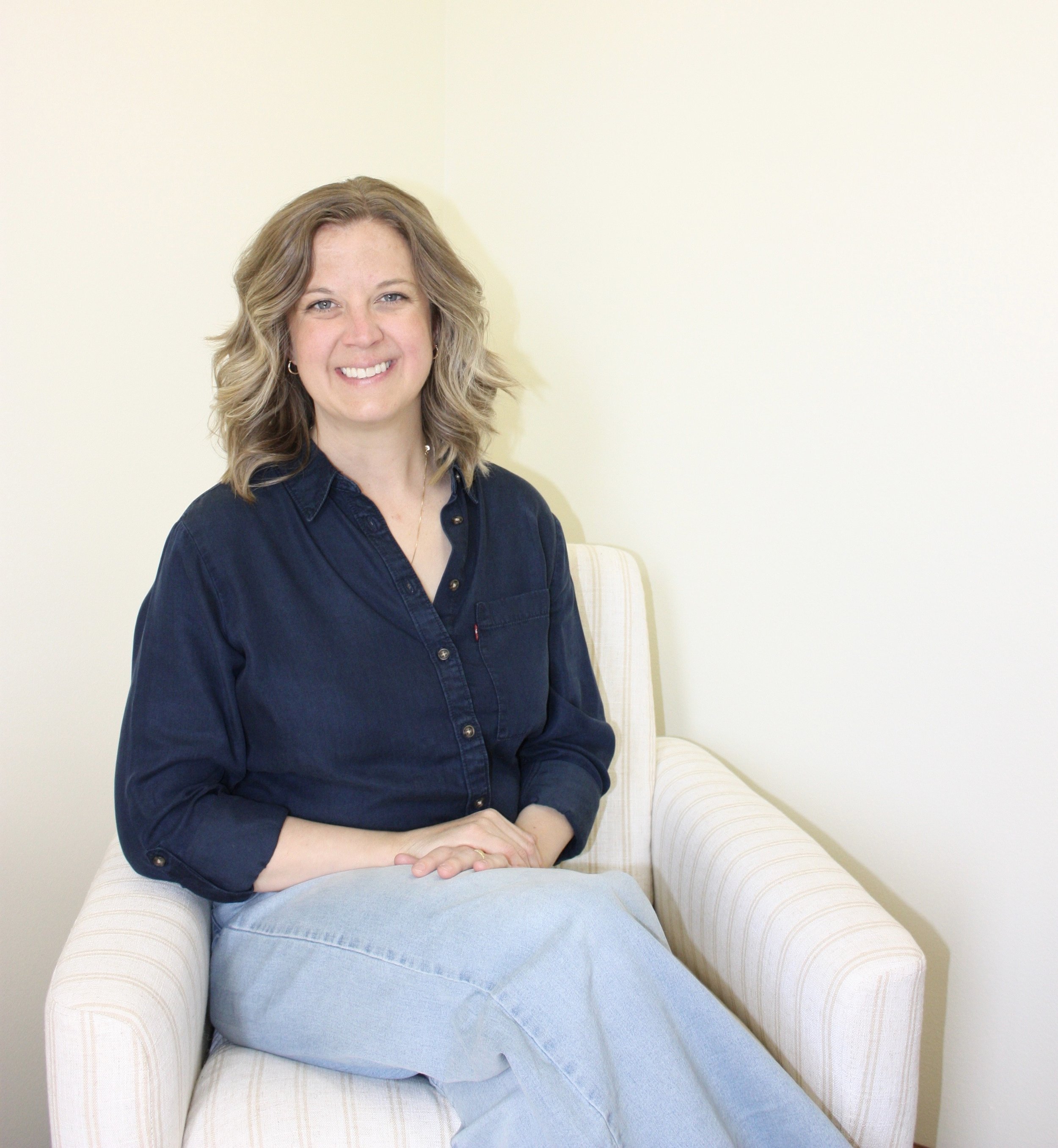 A woman with shoulder-length wavy blonde hair, wearing a dark blue shirt, smiling while sitting on a light-colored, striped armchair with a matching cushion, against an off-white wall.