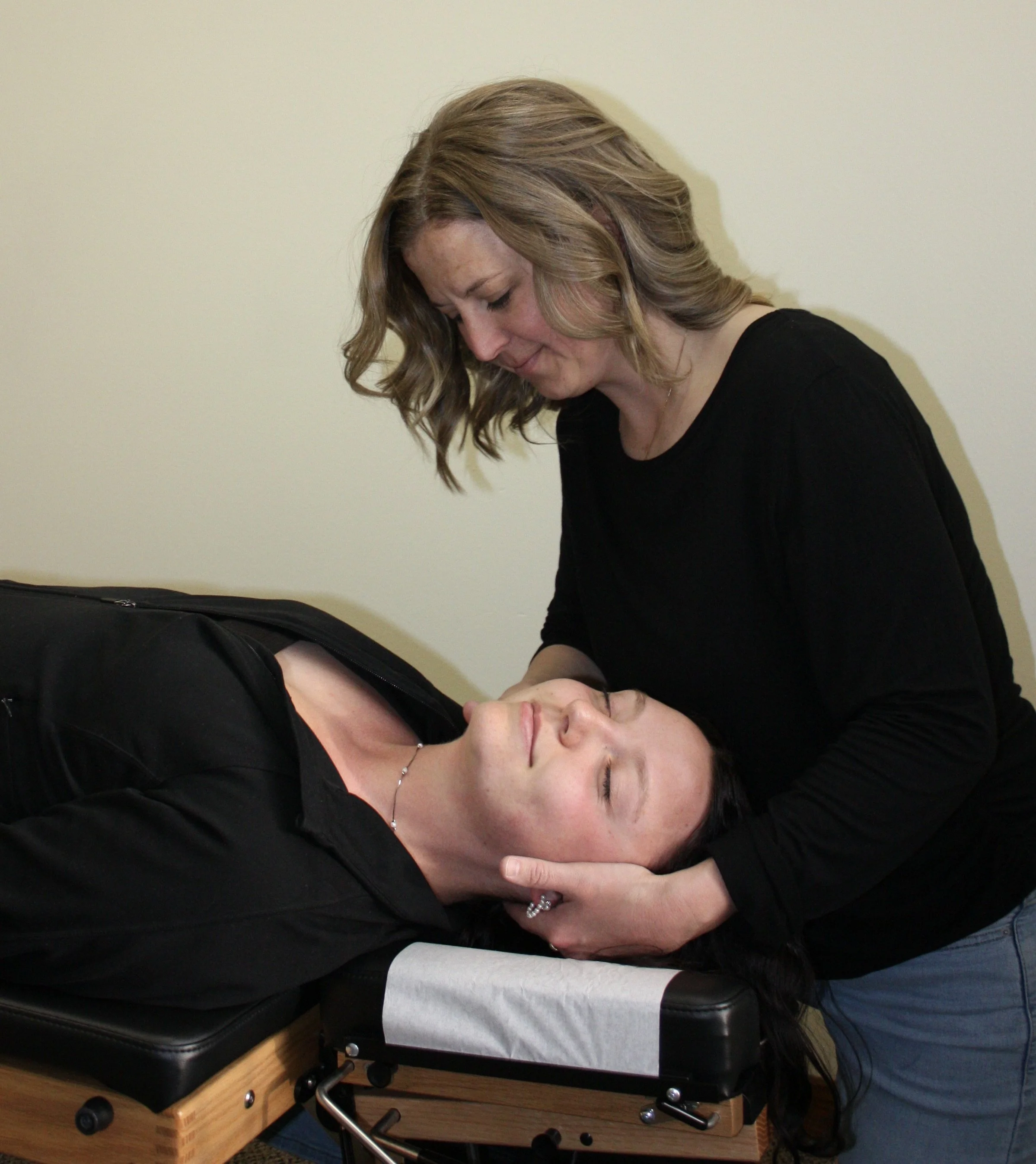 A woman with long dark hair and earrings lying on her side on a massage table with a gray sheet, her eyes closed. A woman with light brown hair styled in waves, wearing a black shirt, leaning over her, supporting her head with her hand, and smiling gently.