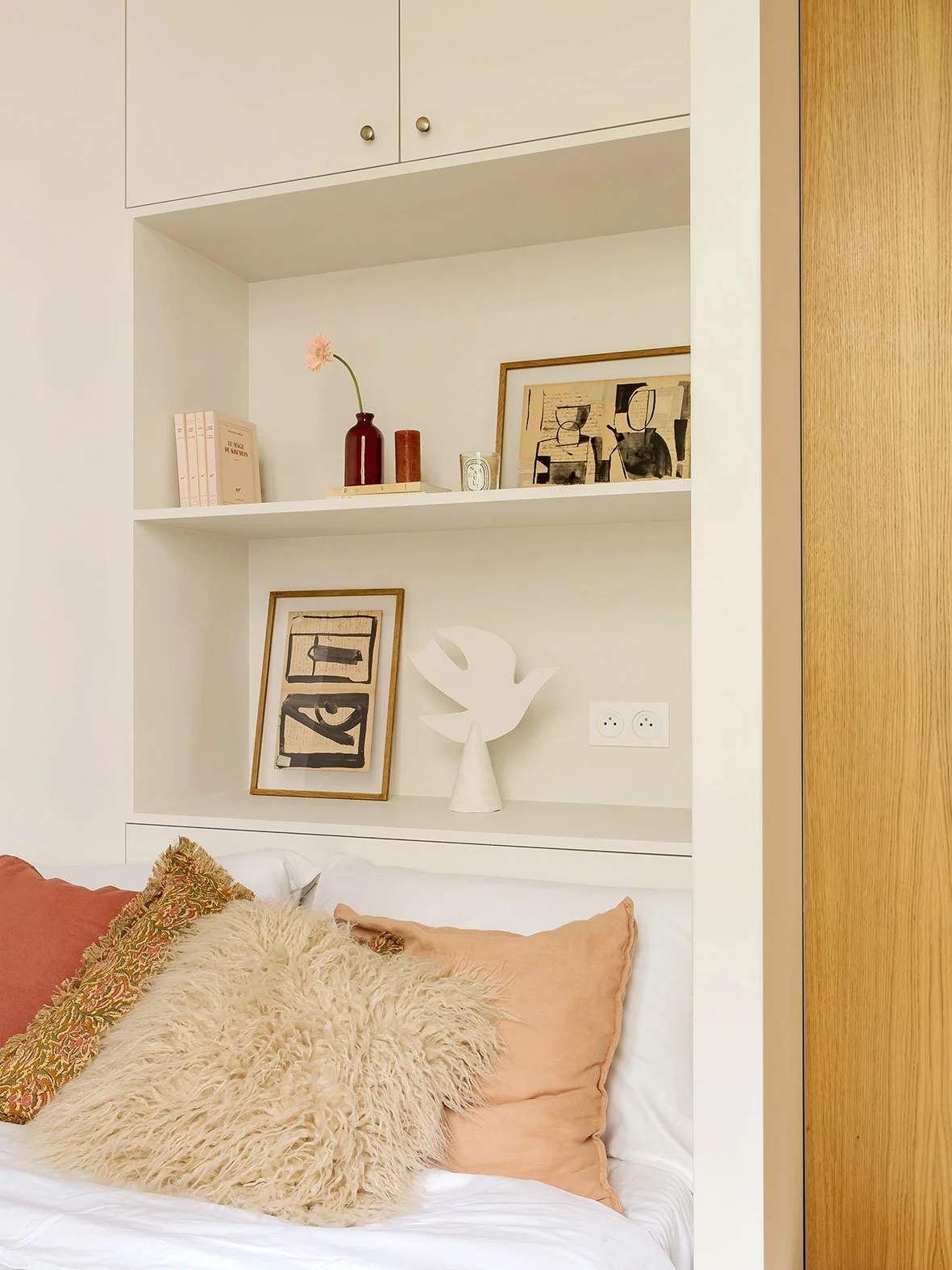 Intérieur d'une chambre avec des coussins en peluche et en tissu sur un lit, étagère blanche derrière avec des décorations, tableaux et sculptures, et un mur en bois à droite.