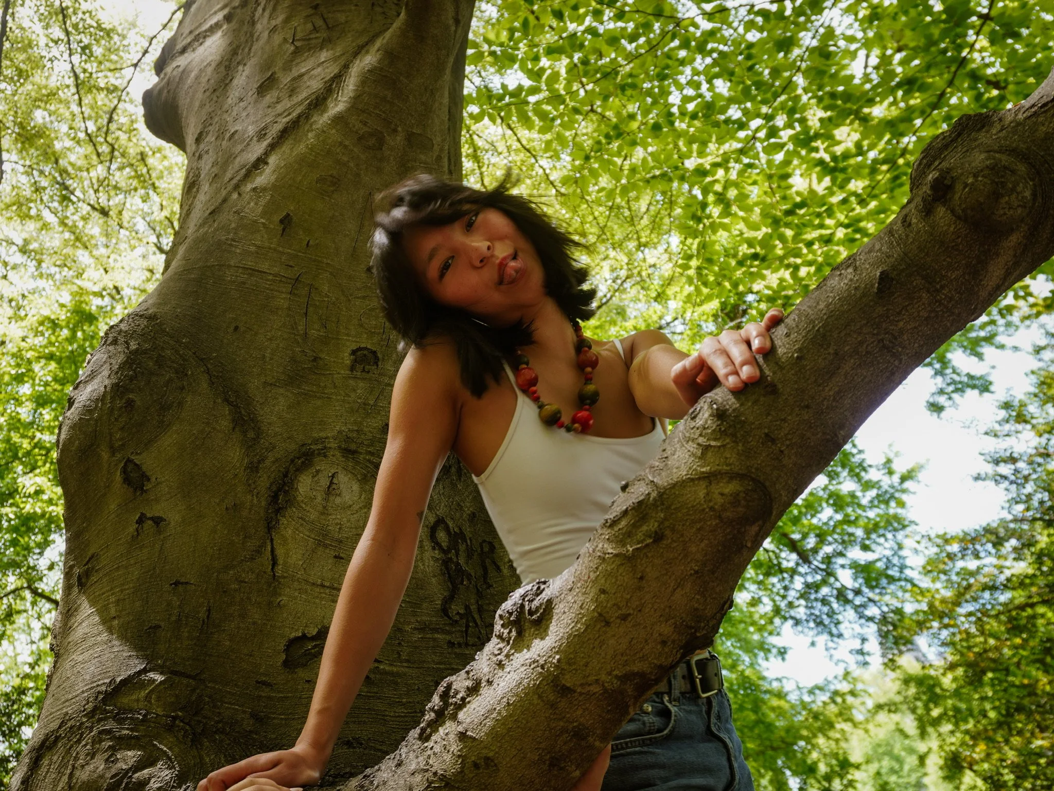 A young woman with dark hair and a colorful beaded necklace in a white tank top climbing a large tree with green leaves, sticking her tongue out playfully.