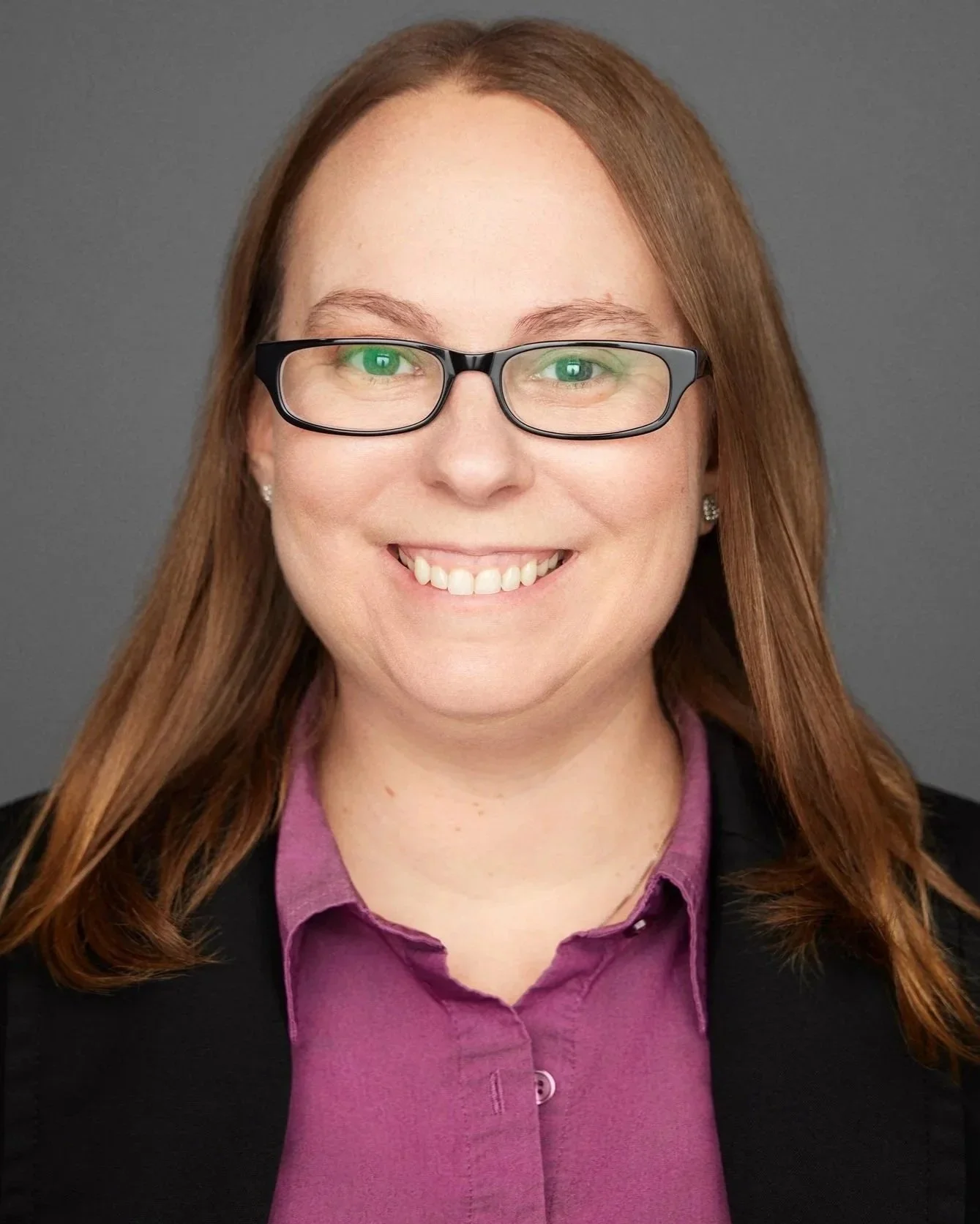 A woman with red hair wearing glasses, a black blazer, and a purple shirt, smiling at the camera against a gray background.