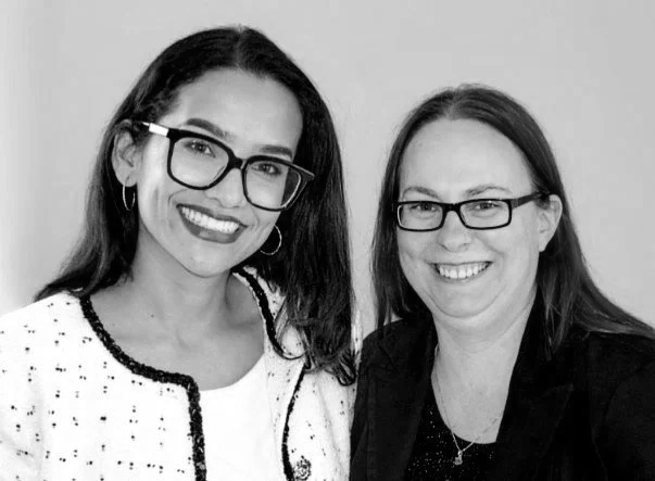 Two women with glasses smiling and standing close together.