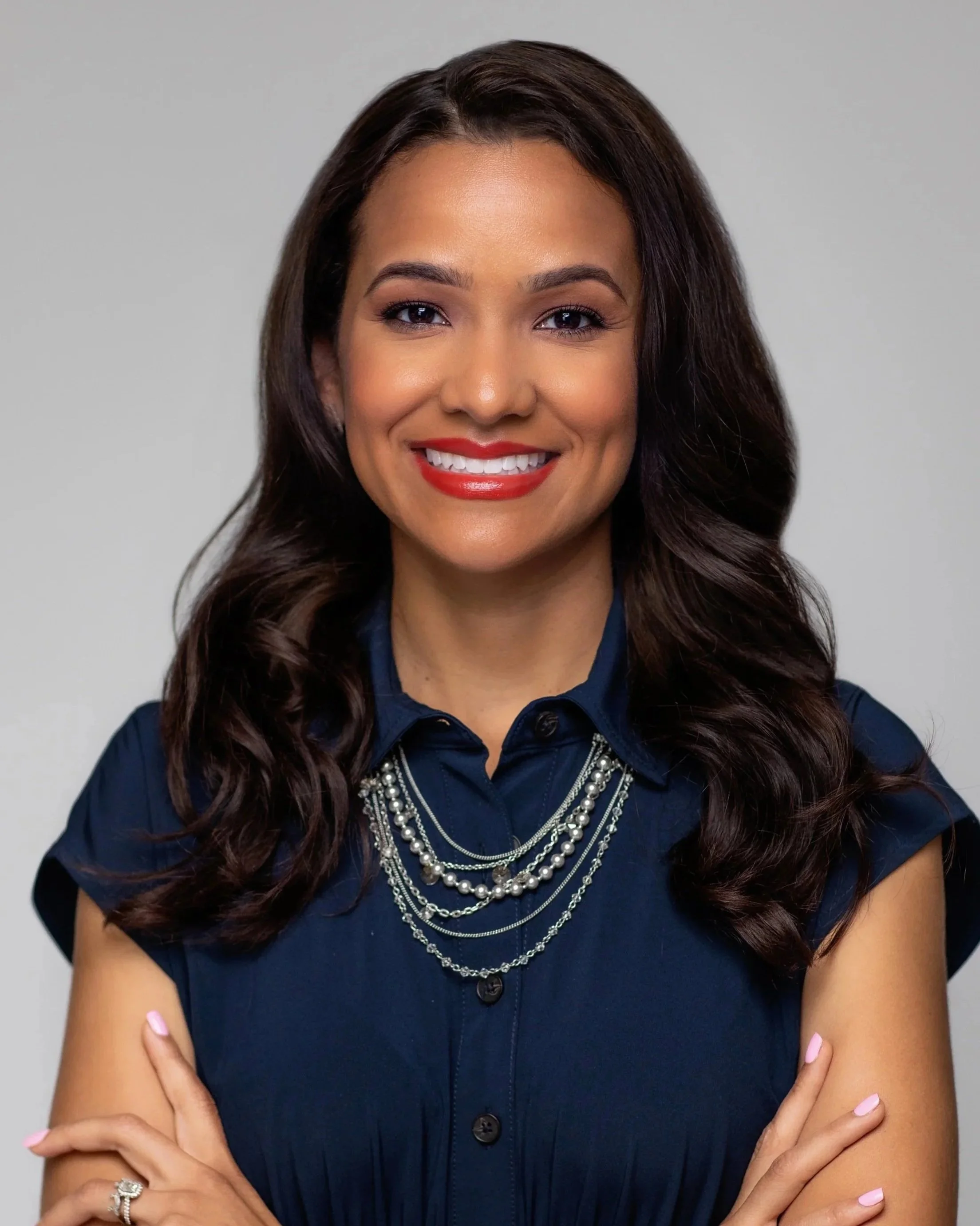 Close-up of a woman with long dark hair wearing a navy blue button-up shirt and multiple silver necklaces, smiling with crossed arms against a plain gray background.
