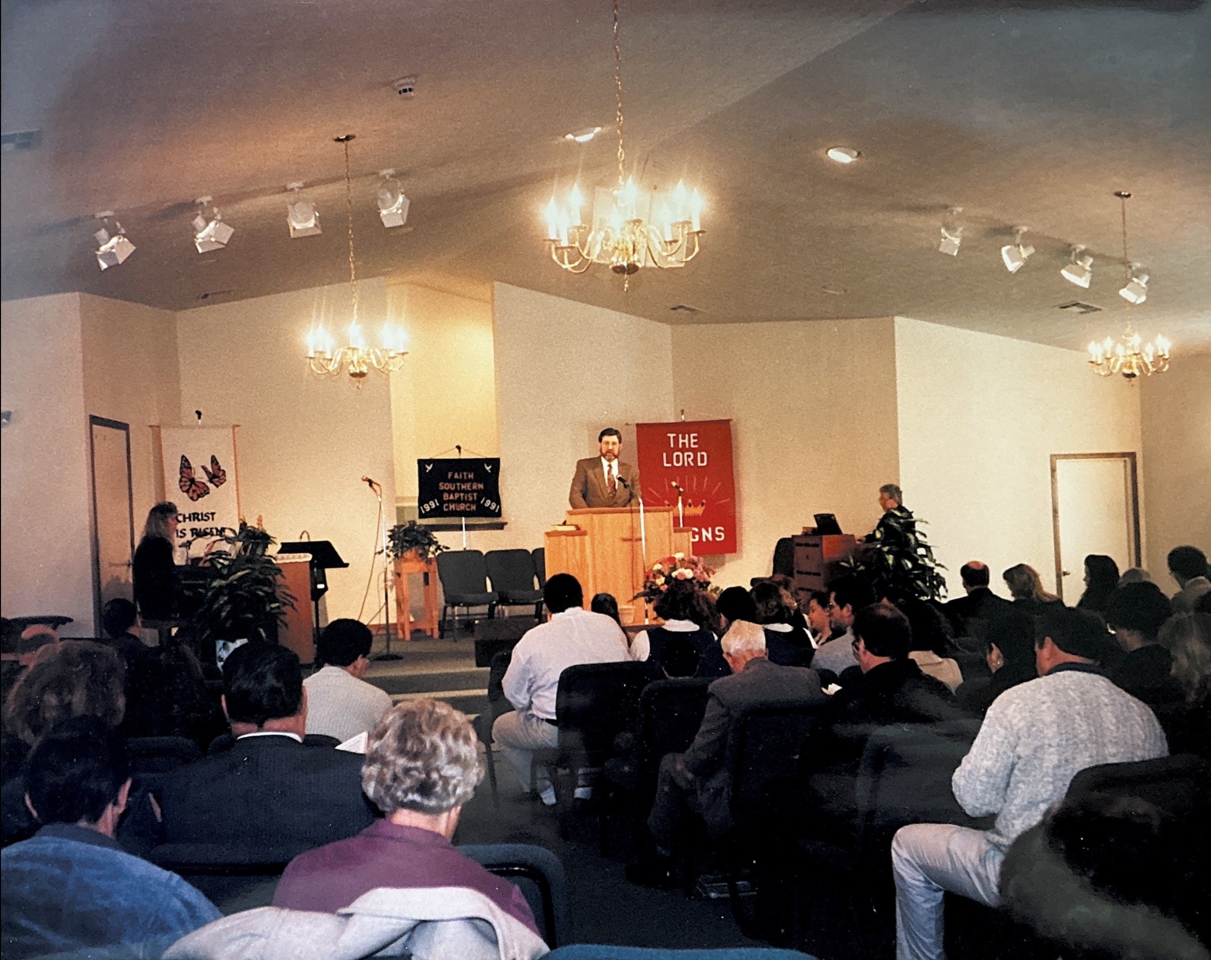 Audience seated in a room watching a man giving a presentation, with a woman playing guitar and a person at a music stand in the background.