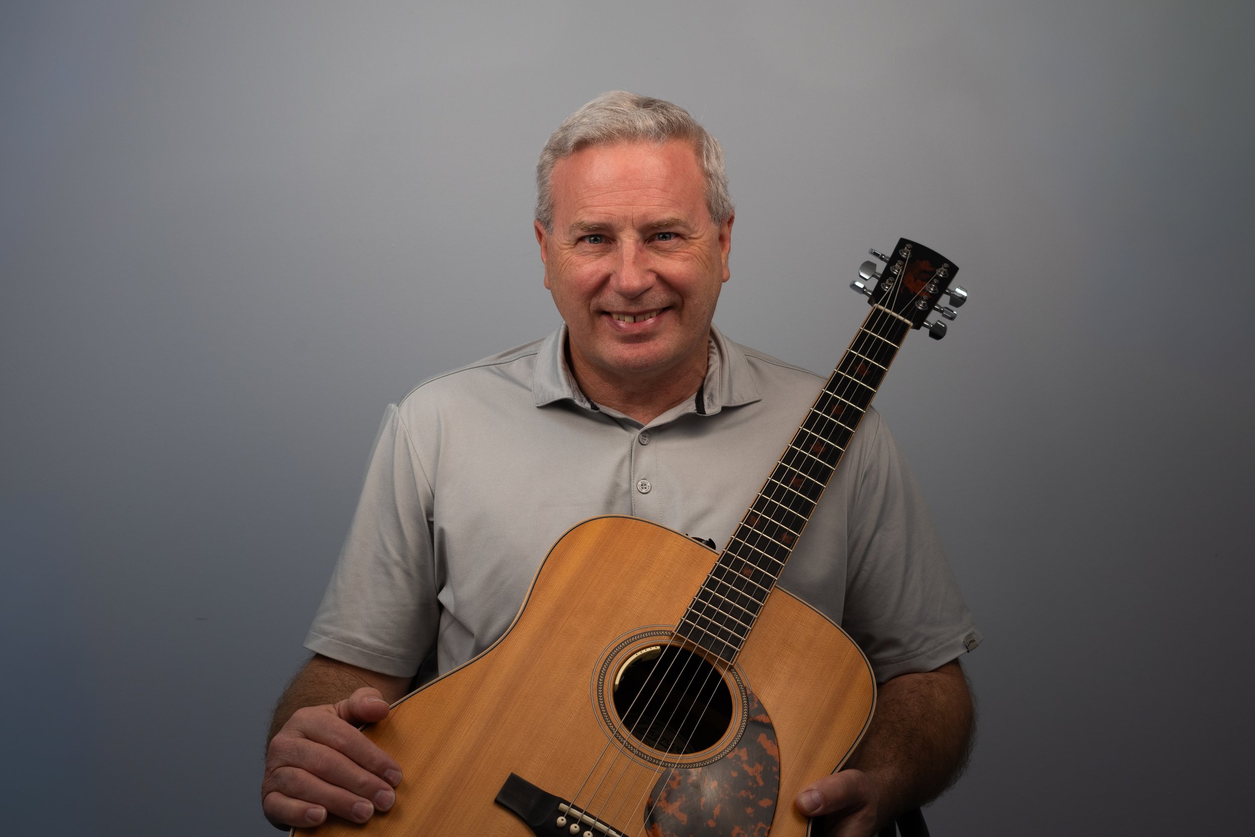 A man with gray hair smiling while holding an acoustic guitar against a plain gray background.