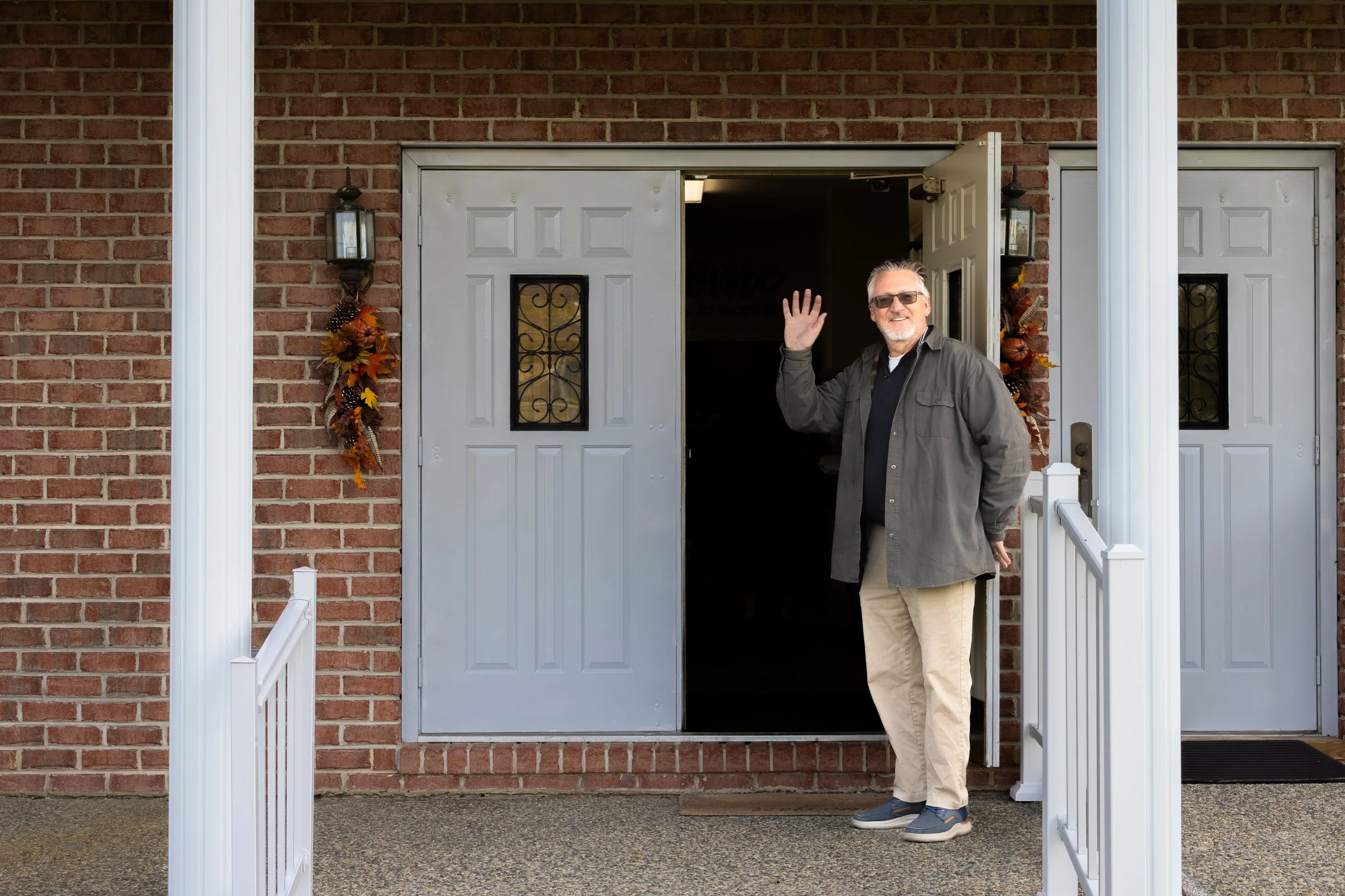 A smiling man with gray hair, wearing sunglasses, a gray jacket, and beige pants, standing at a doorway of a brick building, waving at the camera.