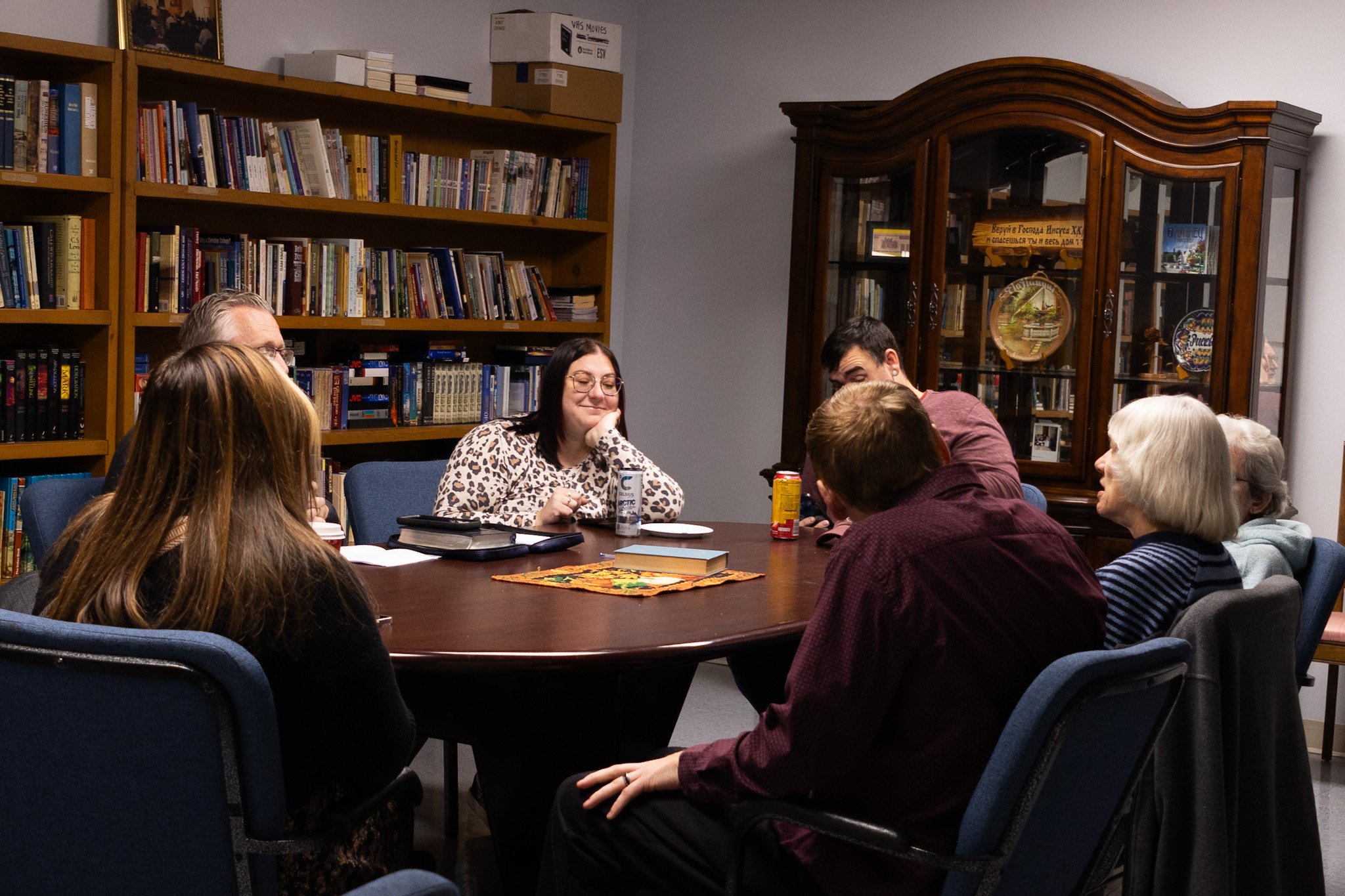 Group of people sitting around a large wooden table in a library, engaging in conversation, with shelves filled with books and framed photos in the background.