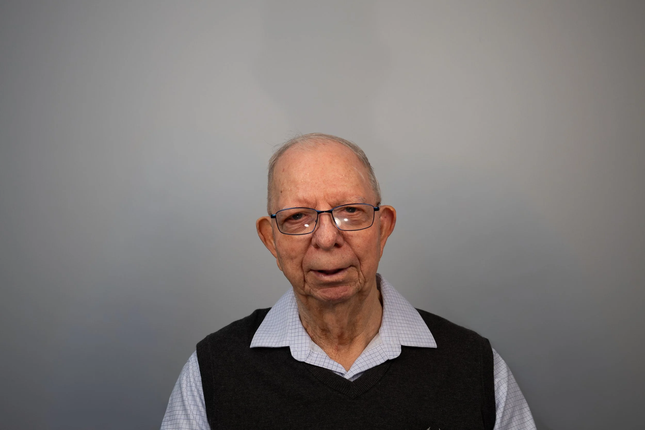 An elderly man with glasses, wearing a collared shirt and a sweater vest, standing against a plain grey background.
