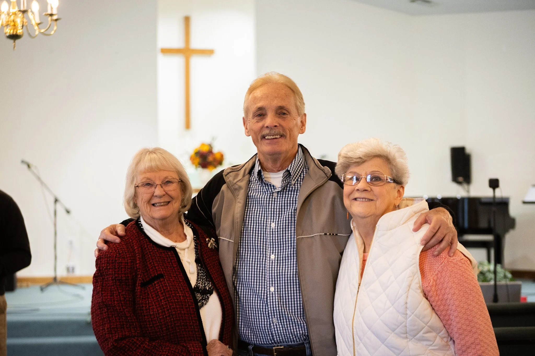 Three elderly individuals, two women and a man, posing together inside a church with a large wooden cross on the wall behind them. They are smiling and hugging each other.