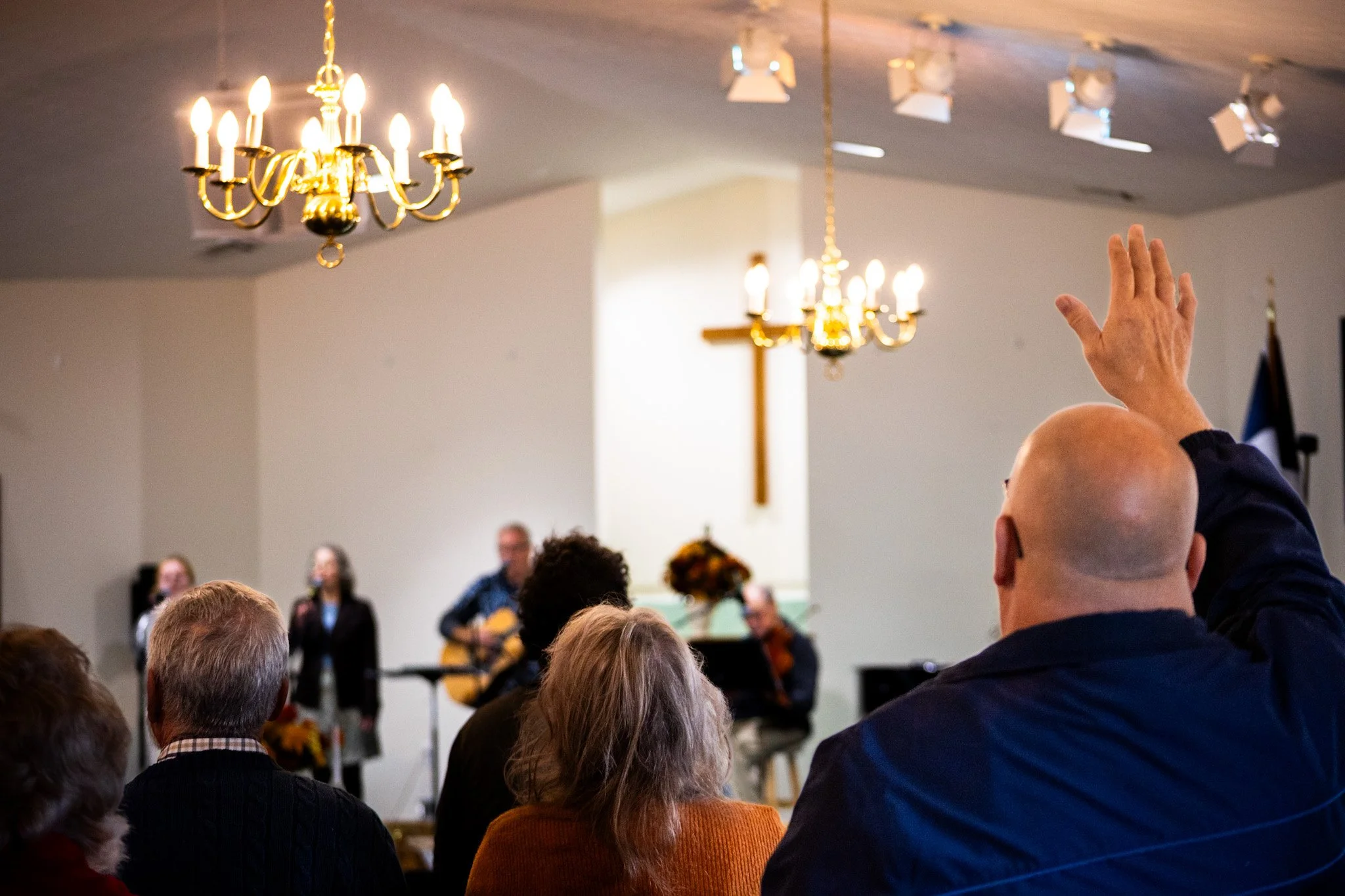 People gathered in a church or meeting hall, with some raising their hands, a person in the background playing guitar, a cross on the wall, and chandeliers hanging from the ceiling.