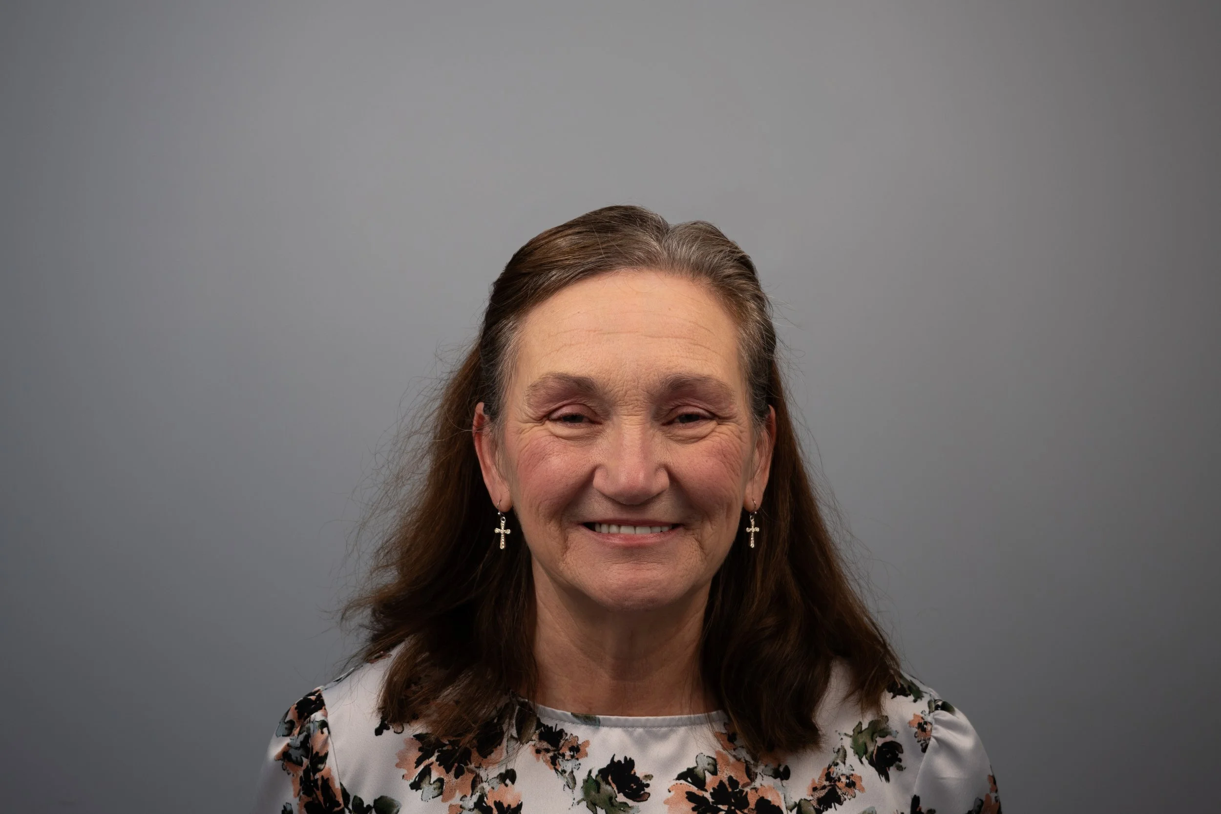 A smiling middle-aged woman with long brown hair and cross earrings, wearing a white floral blouse, standing in front of a plain gray background.