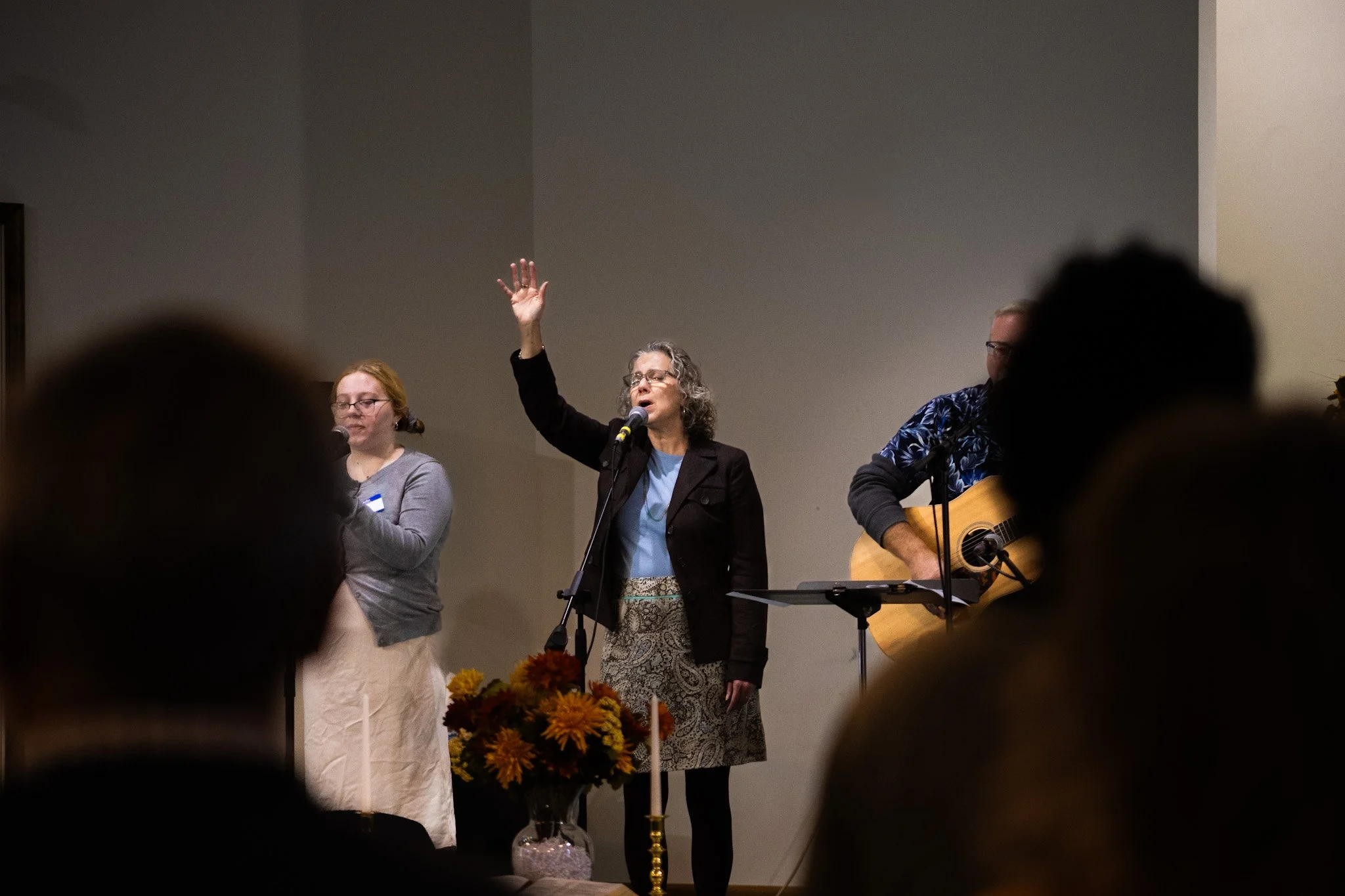 A woman with gray curly hair and glasses raising her hand and speaking into a microphone, accompanied by a woman on her left and a man on her right, playing guitar, in a room with a plain wall in the background.