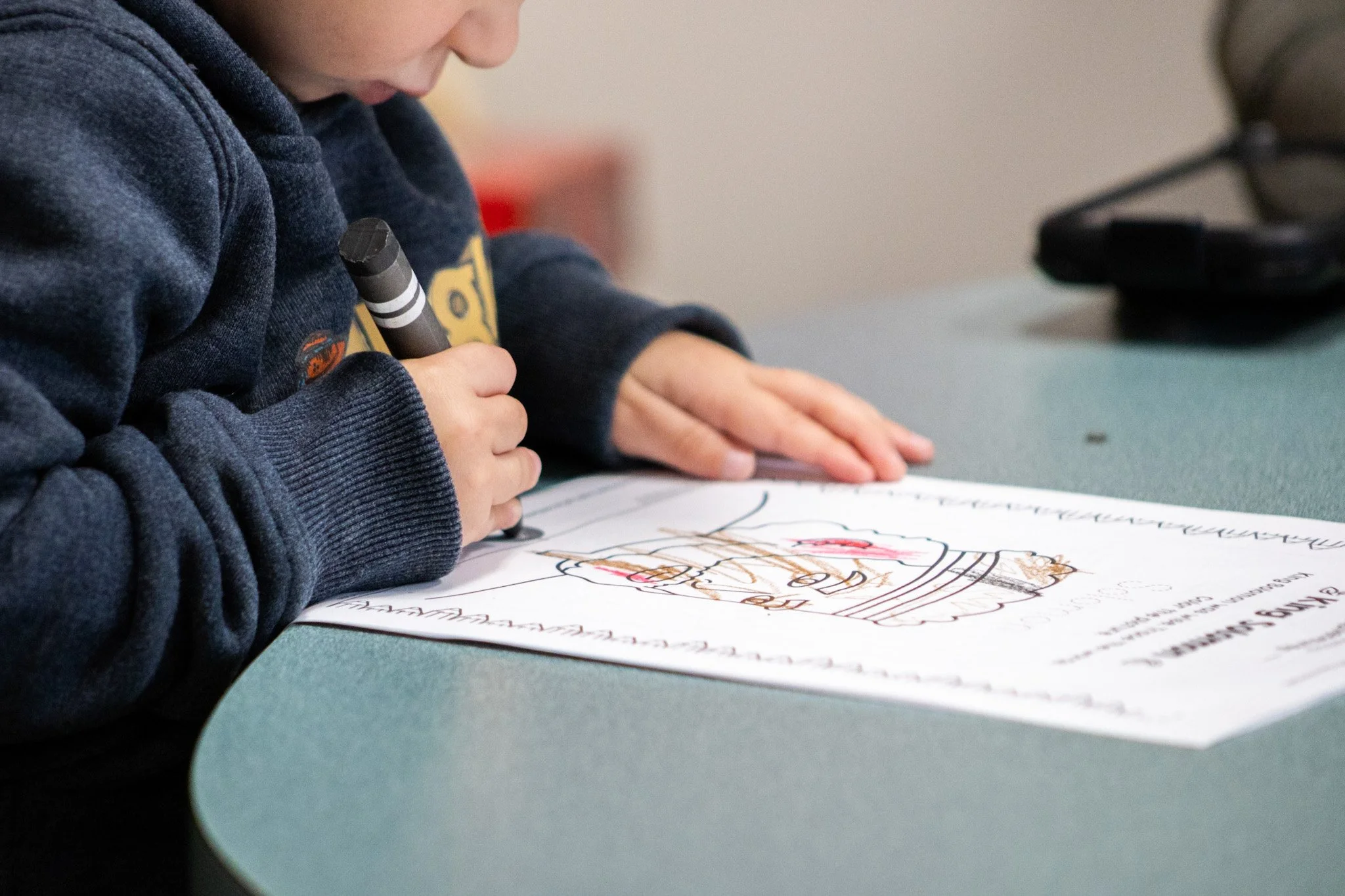 A young boy in a blue hoodie drawing or coloring on a worksheet with a black marker. The worksheet appears to have an illustration with some handwritten text. The boy is seated at a table covered with a light-colored surface, with a black device visible in the background.