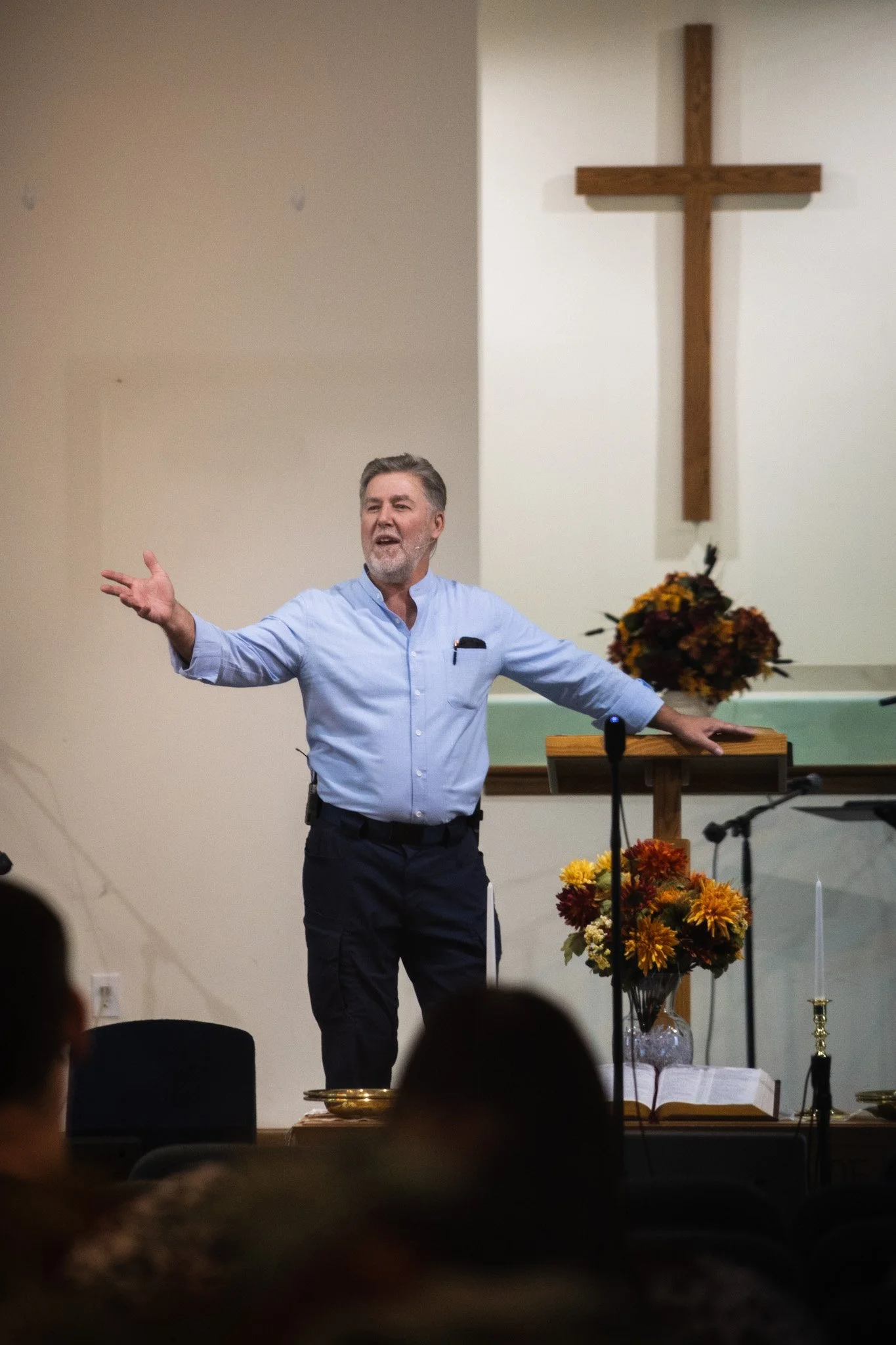 A man with gray hair and beard wearing a light blue shirt is speaking or giving a presentation in a church or religious setting. Behind him is a large wooden cross on a white wall and a table with a bouquet of autumn-colored flowers.