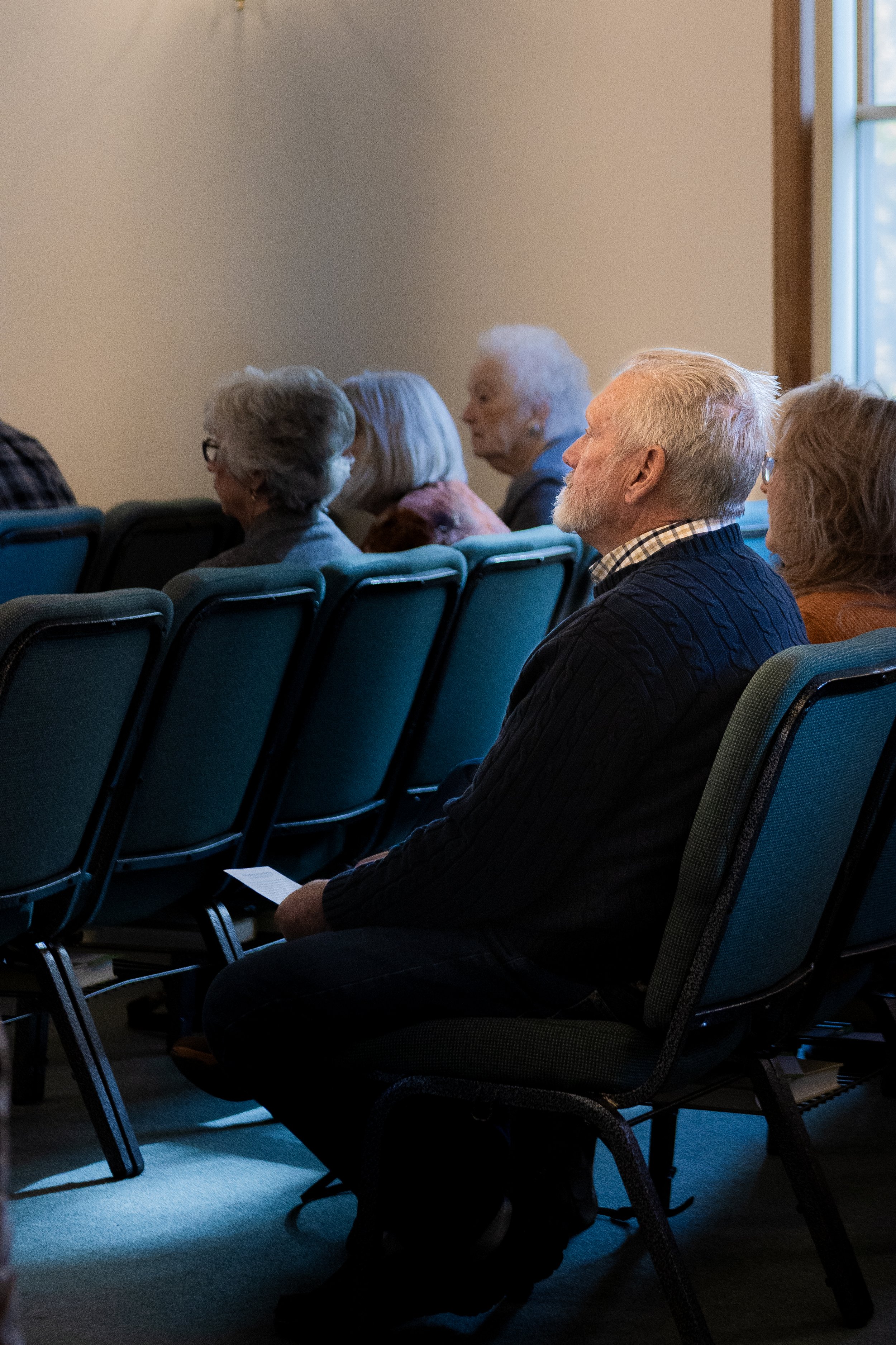 A group of elderly people seated in chairs attending a meeting or presentation in a room with natural light coming through a window.