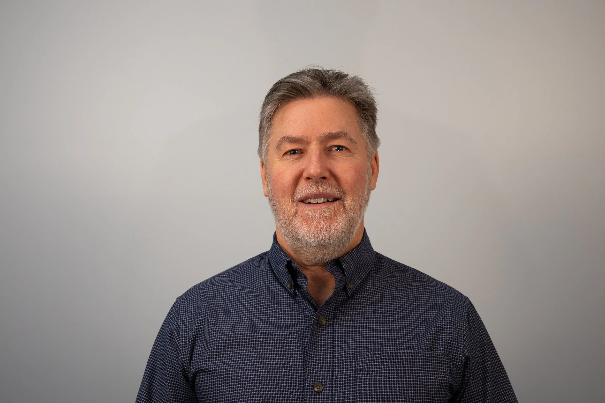 Portrait of a middle-aged man with gray hair and beard wearing a dark checkered shirt against a plain light gray background.