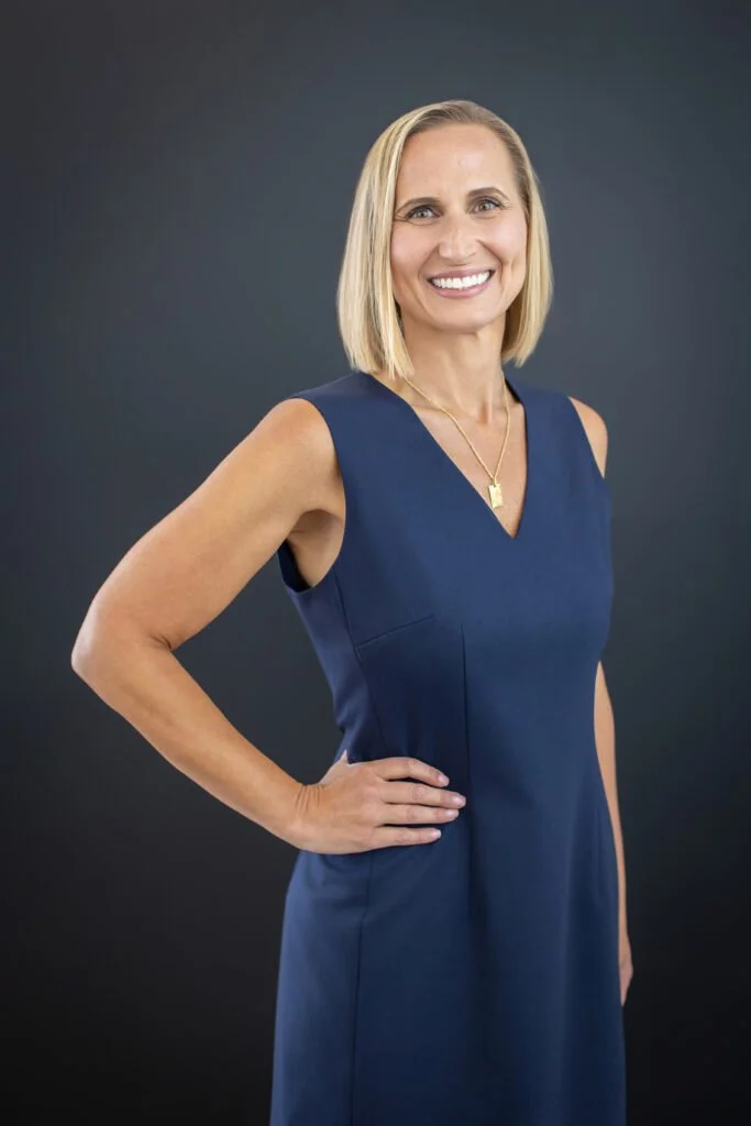 A smiling woman with blonde hair wearing a sleeveless navy blue dress and a gold necklace, standing against a dark gray background.