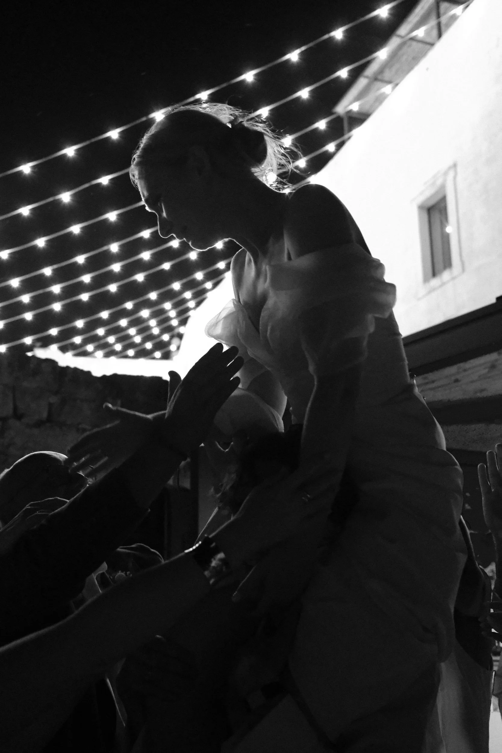 Bride lifted above the wedding crowd under festoon lights, hands reaching up