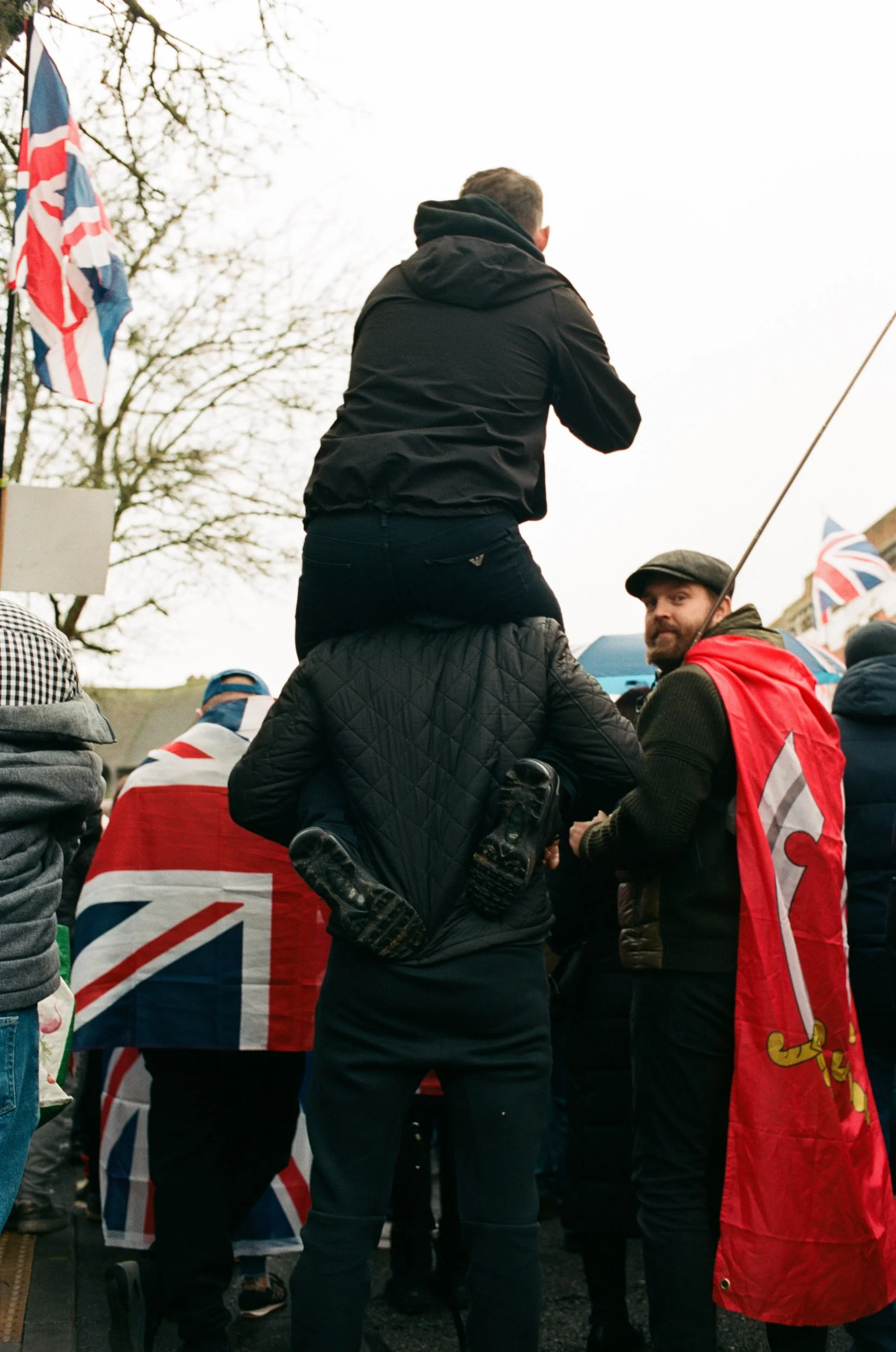Man carried on shoulders at a march while a caped man turns to face the camera