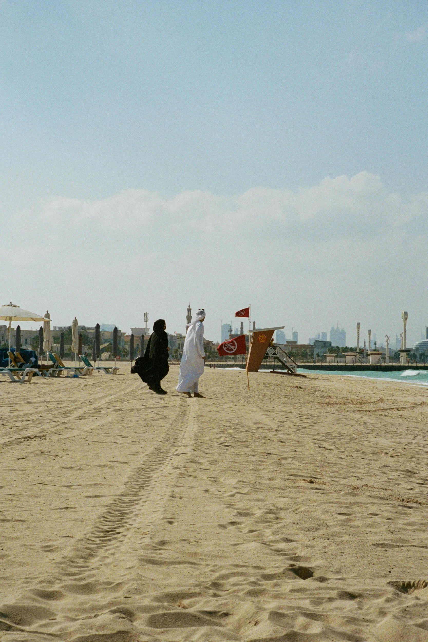 An Emirati couple walk past red no-swim flags, minaret and skyline in the haze