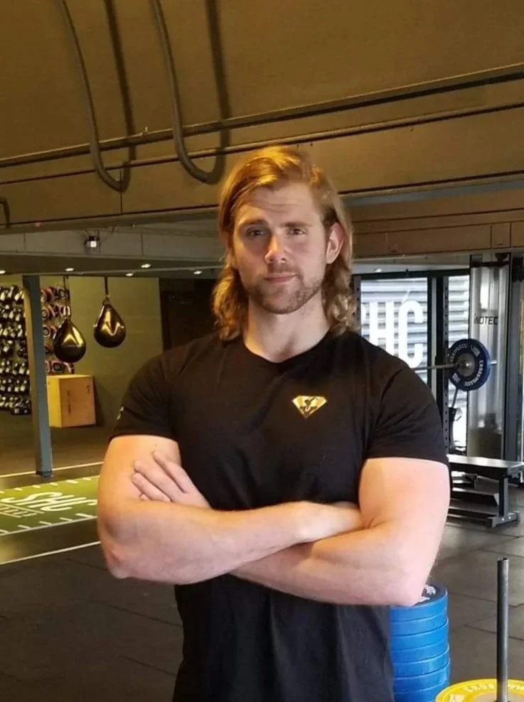 A man with long blonde hair and a beard standing with arms crossed in a gym, wearing a black T-shirt with a yellow logo, fitness equipment visible in the background.