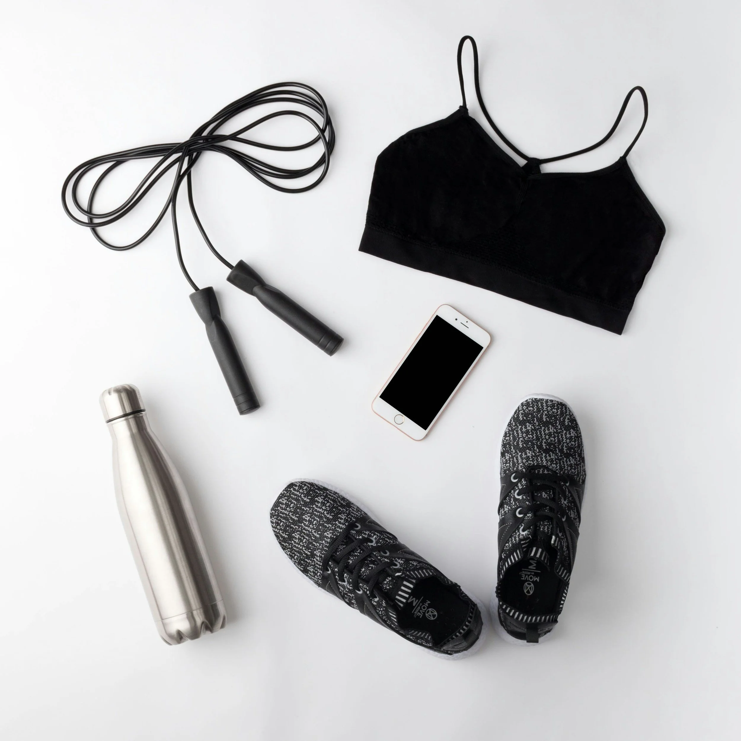 A black face mask, a jump rope with black handles and black rope, a silver water bottle, a white smartphone with a gold frame, a pair of black and white patterned athletic shoes, and a person's legs in black and white running shoes are laid out on a white surface.