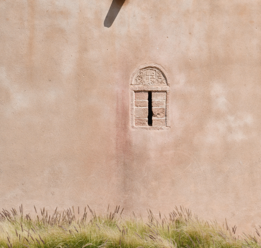 Mur en pierre avec une petite fenêtre en arc et barre verticale, dans un mur en plâtre rose clair, avec de l'herbe au premier plan.