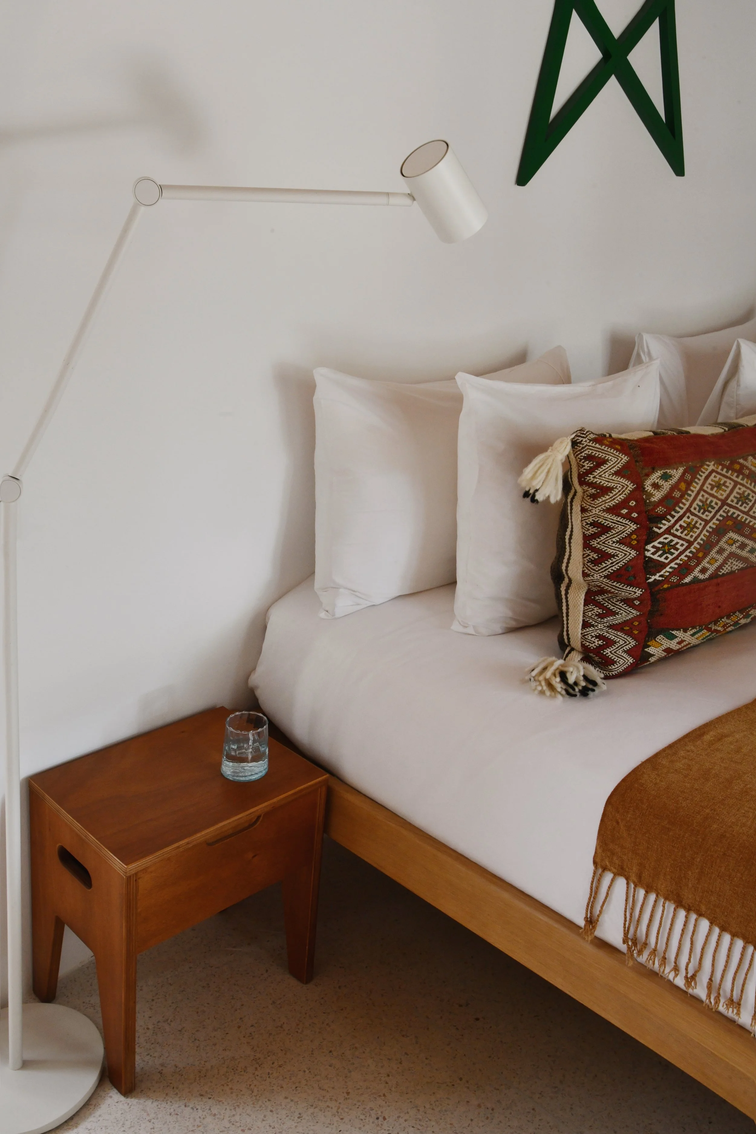 Coin de chambre avec lit, deux oreillers, couverture orange, lampe de chevet et verre d'eau sur une petite table en bois.