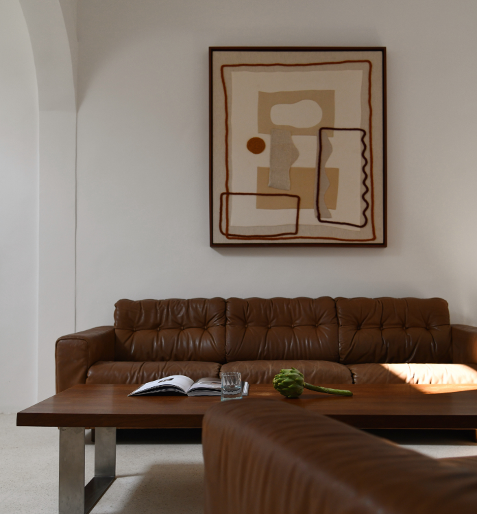Un salon avec un canapé en cuir marron, une table en bois avec un livre ouvert, un verre d'eau et une sculpture verte, tableau abstrait accroché au mur blanc en arrière-plan.